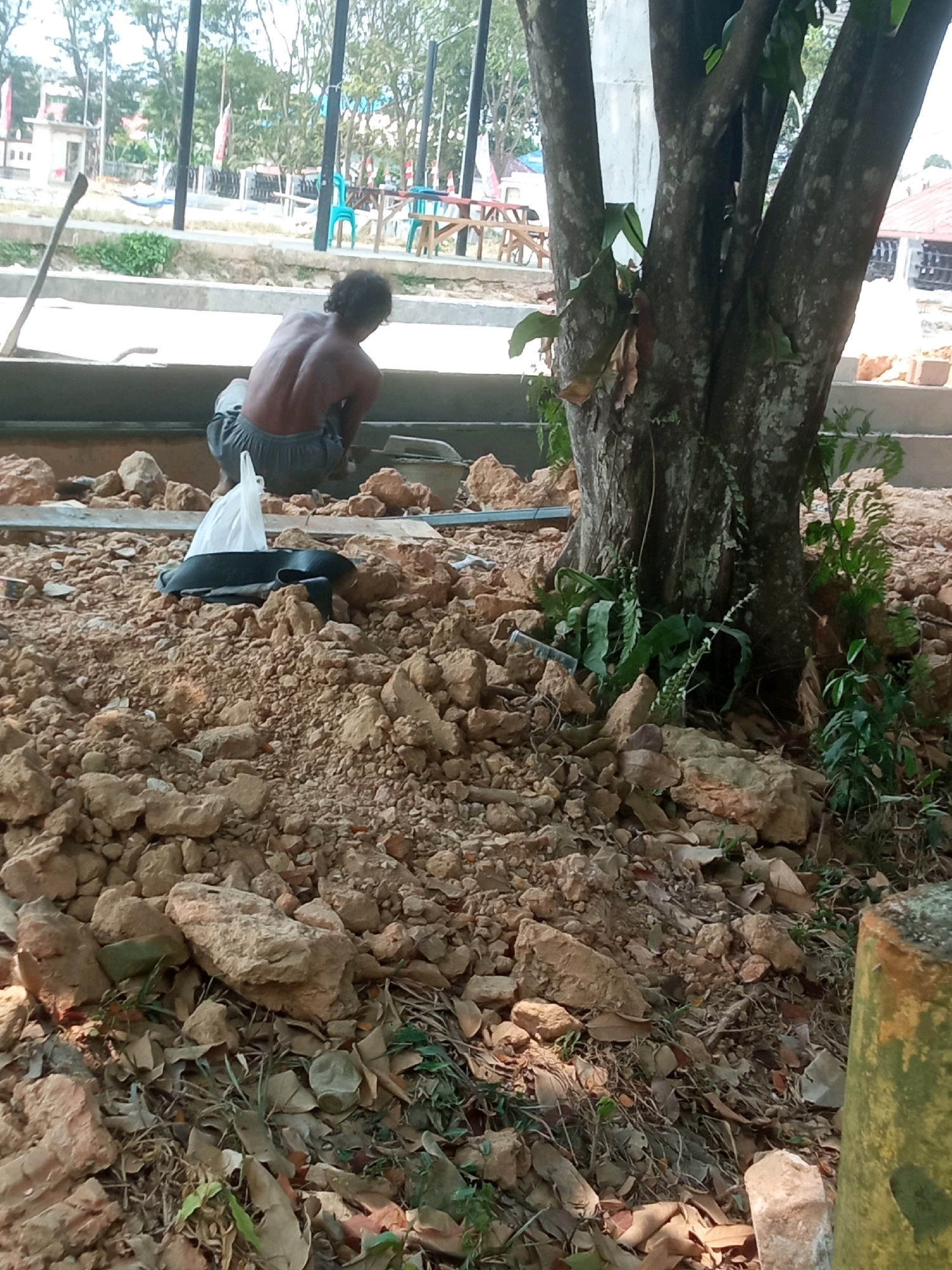 The image depicts a construction site in Bontang, Indonesia, with a bare-chested man kneeling on the ground, possibly engaged in construction work. He is wearing grey shorts and appears to be focused on something in front of him. The foreground is dominated by piles of rocks and soil, indicating ongoing excavation or building activities. In the background, a large tree trunk is visible, with some vegetation growing around its base. Beyond that, there are hints of a park-like area with picnic tables and chairs, suggesting the construction is taking place in a public space or a park. The overall scene suggests an outdoor, daytime environment with a focus on manual labor and construction.