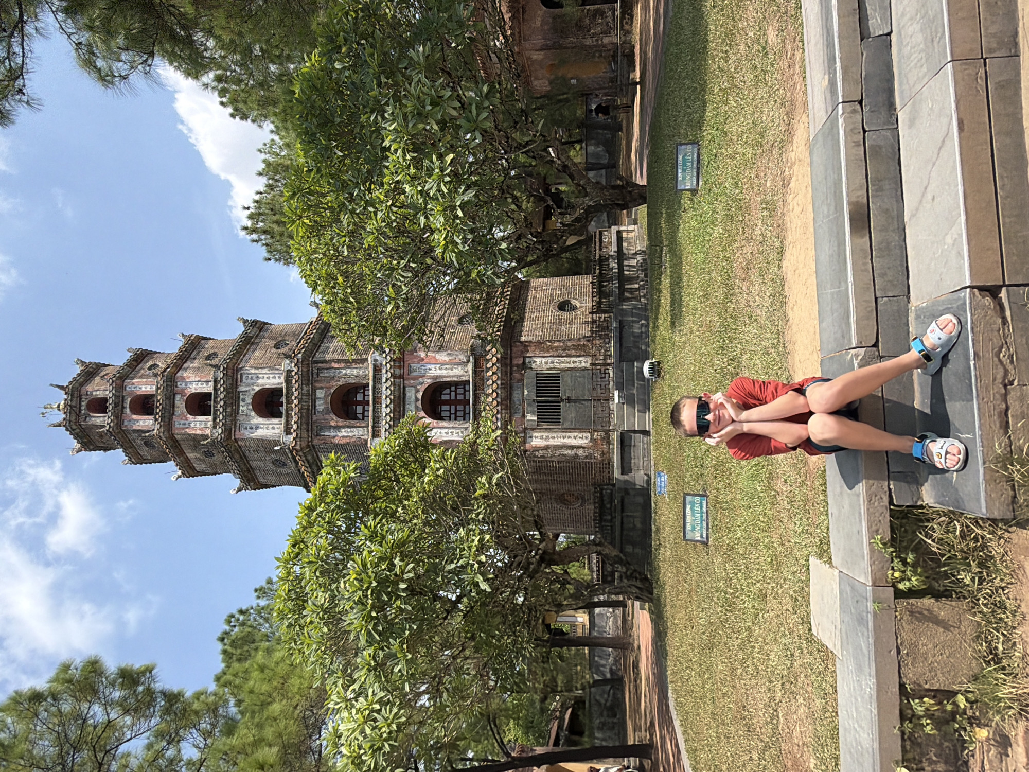 A young boy, wearing a red t-shirt, dark shorts, sunglasses, and sandals, sits on a stone ledge in front of a multi-tiered pagoda. He has his hands propping up his chin and is looking at the camera. The pagoda, made of brick and adorned with decorative elements, has several levels and arched windows. Lush green trees flank the pagoda on either side. In the background, there are other structures and more trees, suggesting a park or historical site. The ground is covered in grass and a pathway. Two small signs are visible in the grass in front of the pagoda. The sky above is blue with scattered clouds. The scene appears to be in Kim Long Ward, Vietnam, indicated by the architectural style and vegetation.