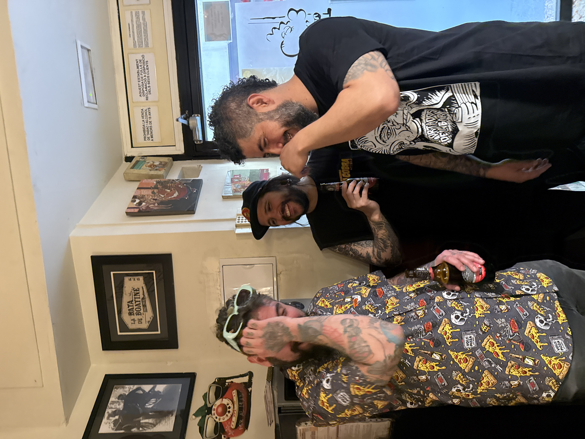 Three tattooed men are indoors, appearing jovial. One is covering his eyes with his hand, wearing light green sunglasses on his forehead and a button-down shirt with a pizza, skull, and other graphic pattern. He holds a brown beer bottle. Another man is wearing a black baseball cap and holding a brown object. The third man, wearing a black T-shirt with a graphic, is touching the second man's face.
On the walls are framed prints of art and posters with messages in both Catalan and Spanish. One poster has text that includes "PROHIBIDA LA VENDA DE TABAC I ALCOHOL A MENORS DE 18 ANYS". There is a red and white telephone, as well as a sign that reads "LA BATA DE BOATINE". The graphic on the t-shirt says "URTADES DEL BAR". One of the posters mentions that the establishment has claim forms at the disposal of clients. Given the Catalan language, the location is most likely in the city of Barcelona, Spain.