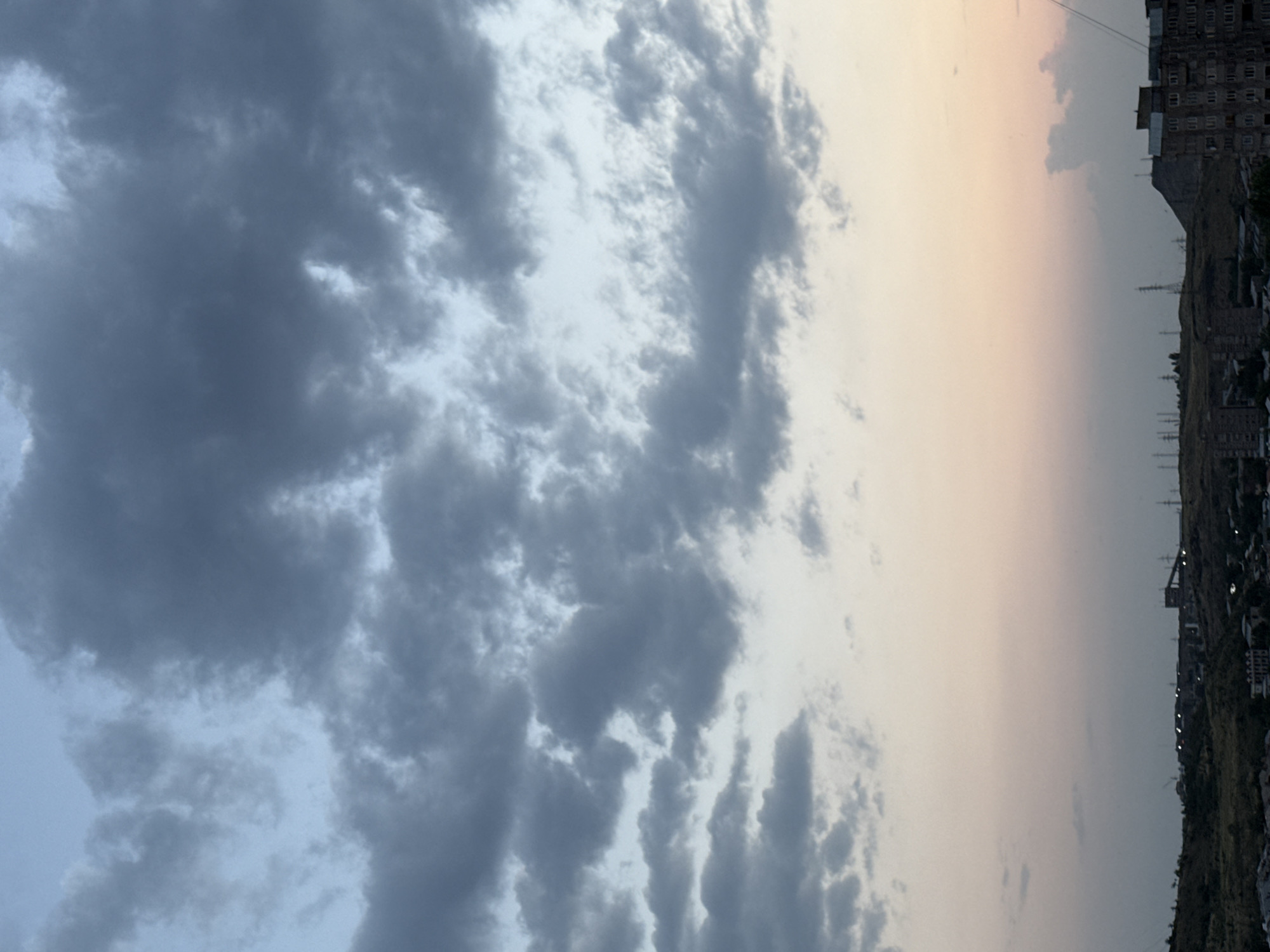 This image captures a landscape view of Abovyan, Armenia, likely taken at dusk. The sky is filled with dramatic, dark gray clouds interspersed with patches of lighter blue and a soft pinkish-orange hue near the horizon, suggesting the transition from day to night.
Below the sky, a rolling hilly terrain is visible, dotted with various structures. In the foreground, on the right, a multi-story apartment building with visible windows and balconies is prominent. Further in the background, along the horizon line, there is a dense collection of industrial or urban buildings, characterized by varied rooflines and some appear to be low-rise structures. Several power lines and transmission towers are scattered across the landscape, indicating an active utility infrastructure. A prominent hill or mountain is visible in the mid-ground, with a dark silhouette. The overall impression is one of an urban or semi-urban environment against a vast, cloudy sky. No people are discernible in the image.