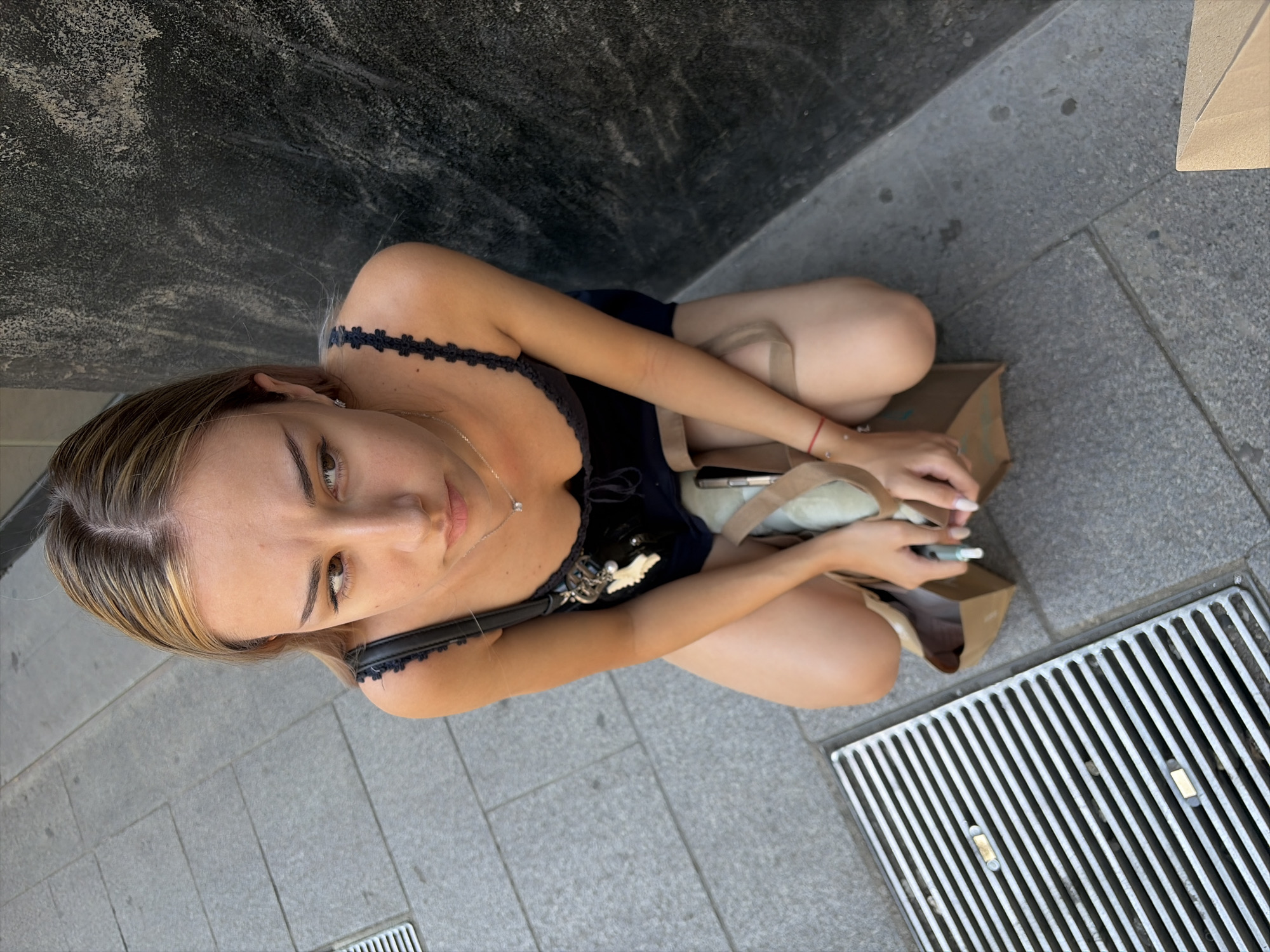 This media file shows a young woman sitting on a sidewalk in Milan, Italy. She is wearing a dark blue top with lace trim and has a delicate silver necklace with a small pendant. A dark choker with a floral pattern is visible around her neck. She is holding a brown paper bag and a slim, cylindrical object, possibly a vape or e-cigarette. The ground is paved with grey tiles, and a metal grate is visible to her right. To her left, a dark, textured wall is present. The lighting suggests it is daytime, likely sunny. The woman is looking directly at the camera with a neutral expression. The scene appears to be outdoors, on a street or public area.
