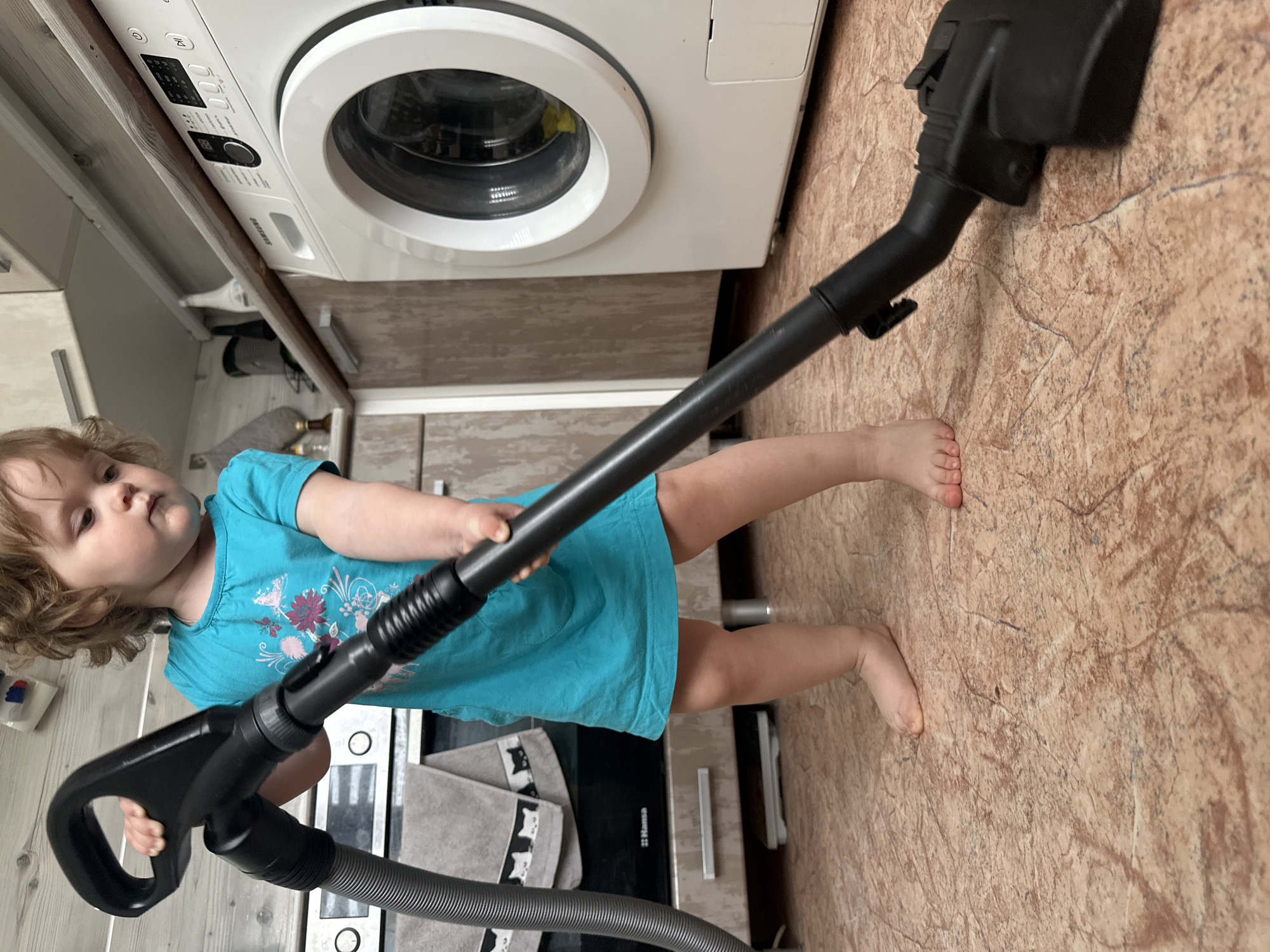 A young child with curly hair is holding a vacuum cleaner in what appears to be a kitchen or utility room. The child is wearing a turquoise t-shirt and is barefoot. The vacuum cleaner has a black wand and a flexible grey hose. Behind the child is a white washing machine and a black stovetop with grey towels decorated with cat faces. The floor has a patterned brown surface. The location is Chaikovsky, Russia.