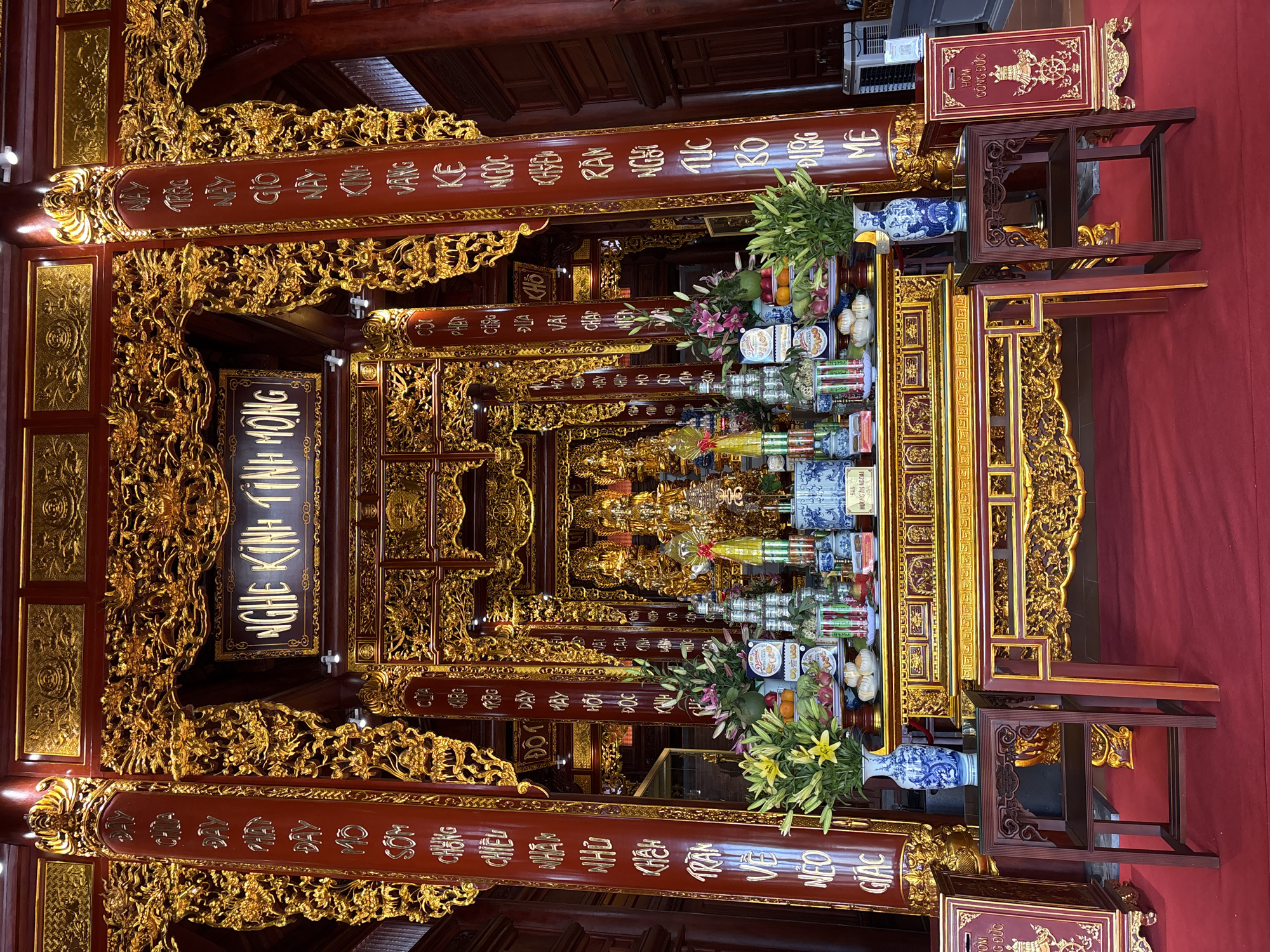 The image shows an ornate altar within a temple, likely in Diên Khánh, Vietnam. The altar is elaborate, with rich red and gold detailing. It is adorned with offerings, including flowers, fruits, and what appear to be canned goods or small packages stacked high. Several vases, including a prominent blue and white porcelain one, are also present. Behind the offerings, a gilded shrine holds statues, possibly of deities or revered figures. The altar is flanked by large, red pillars inscribed with golden Vietnamese characters. The floor is covered in a red carpet, and intricate golden carvings and architectural elements are visible throughout the scene, suggesting a significant religious or cultural site. The overall impression is one of grandeur and devotion.