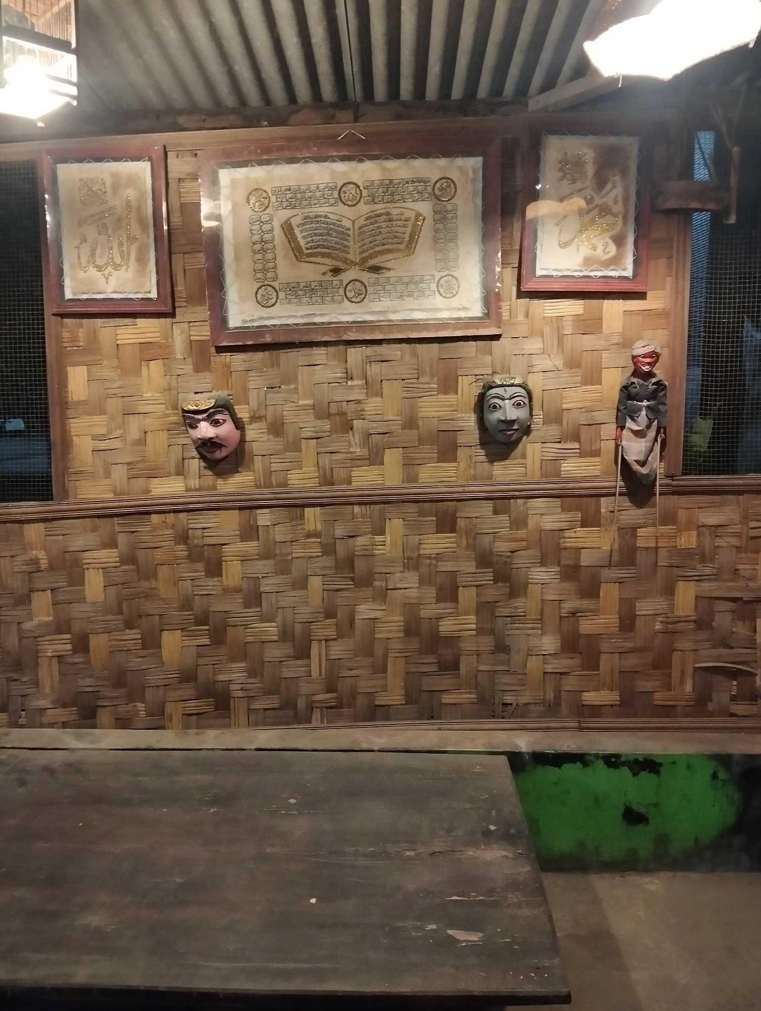 The image depicts an indoor scene, likely a room or a display area, with a dominant woven bamboo wall. Mounted on the wall are three framed artworks, one of which appears to be a representation of an open book with calligraphy. Two decorative masks, possibly of Javanese origin, are also affixed to the wall, along with a puppet figure. A dark wooden table occupies the foreground, obscuring a portion of the lower part of the scene. The lighting suggests an interior setting, possibly with artificial illumination from above. The environment is rustic, with the woven bamboo and the wooden table contributing to a traditional aesthetic. Based on the provided context, the scene is located in Kedungrejo, Indonesia.