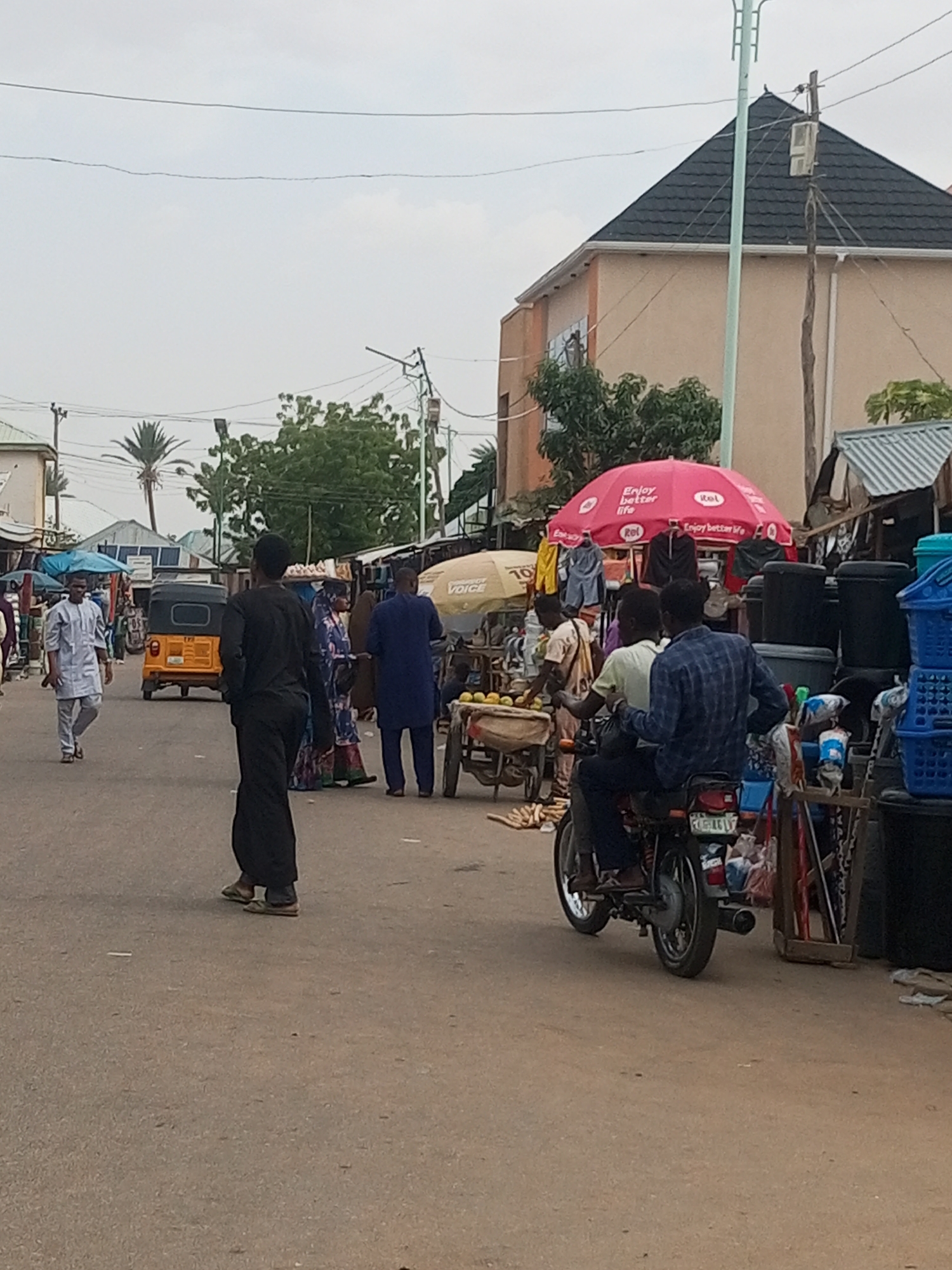 The image shows an outdoor scene at a bustling street market in Gombe, Nigeria. The street is paved and dusty.
**People:** Several people are present, including:
* A man in a white robe walking toward the left of the frame.
* A man in dark clothing is standing on the left, facing away from the camera.
* A woman wearing patterned clothing is seen alongside the man in dark clothing.
* A man in blue clothes.
* A motorcycle carrying two people is positioned on the right side of the street.
**Objects:**
* A yellow auto rickshaw (tuk-tuk) is driving on the street.
* There are stalls selling various goods, including plastic baskets and what appears to be fruit.
* A red umbrella with writing covers one of the stalls.
**Actions:** People are walking, riding, and presumably shopping and selling at the market.
**Environment:** The weather appears to be overcast, as the sky is uniformly bright with no distinct clouds. Buildings of one and two stories stand in the background, with trees dotting the landscape. The power and communication poles are visible above.