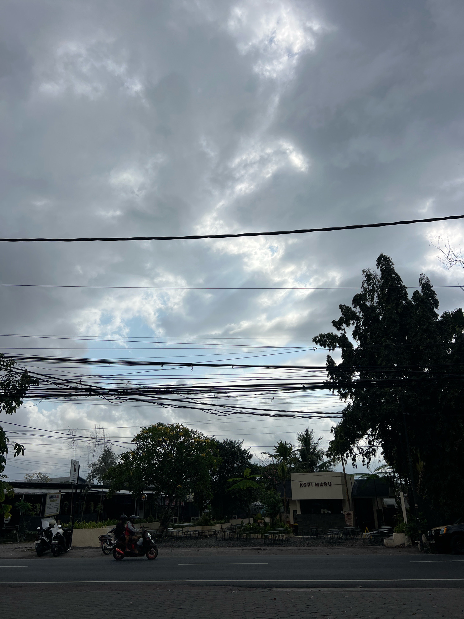 The image depicts a street scene in Denpasar, Indonesia, under a cloudy sky. A two-lane asphalt road occupies the foreground. On the right side of the road, a building with a sign that reads "KOPI MARU" is visible, accompanied by outdoor seating areas and trees. To the left of the building, there are more trees and a few parked motorcycles, one of which is occupied by two people. A few more motorcycles are parked further down the left side of the road. Overhead, a dense network of electrical wires crisscrosses the sky, obscuring much of the view. The overall atmosphere is overcast, suggesting either dawn, dusk, or an impending rain shower.