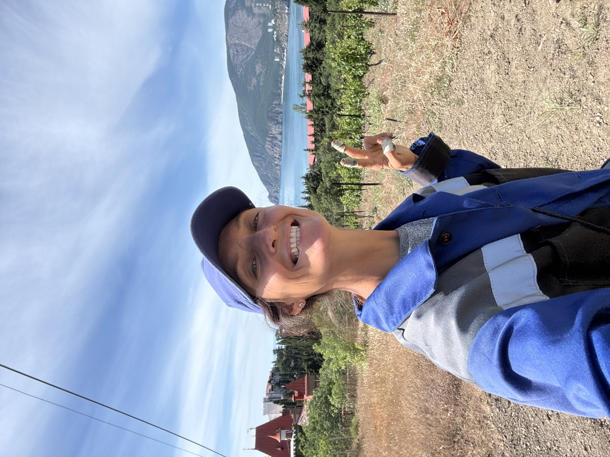 A woman wearing a blue baseball cap and a blue and grey work jacket smiles at the camera while holding up two fingers in a peace sign. Her fingers appear to be wrapped in white tape. She is standing in a dry, grassy area with grapevines in the background. In the distance, there is a body of water, a mountainside, and buildings. The sky is blue with some scattered clouds. The scene is likely in the city of Utios, Ukraine.