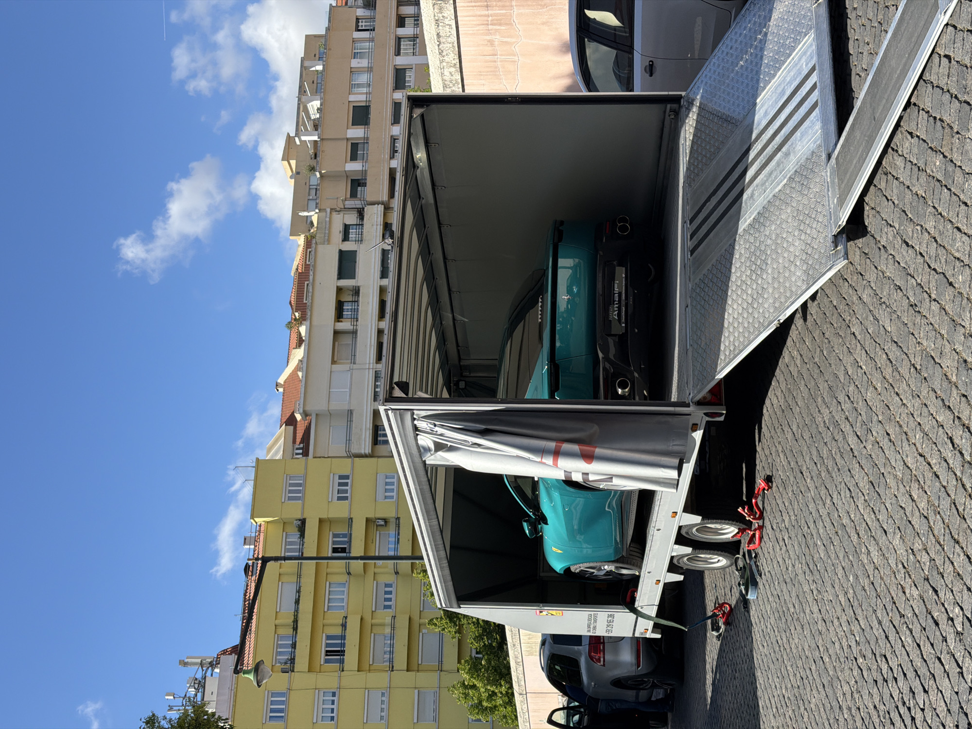 The image shows a car transport trailer with its rear open, revealing two cars inside: a turquoise car in the front and a black car behind it. The trailer is parked on a paved surface, likely a street. The background shows buildings of varying heights and colors, indicative of an urban environment. The sky is blue with scattered clouds. A portion of the trailer's ramp is visible, suggesting the process of loading or unloading. Based on the telephone number (+351 925 165 206), which is a Portuguese mobile number, and the architectural style of the buildings, the location is identified as Lisbon, Portugal. No people are clearly visible.