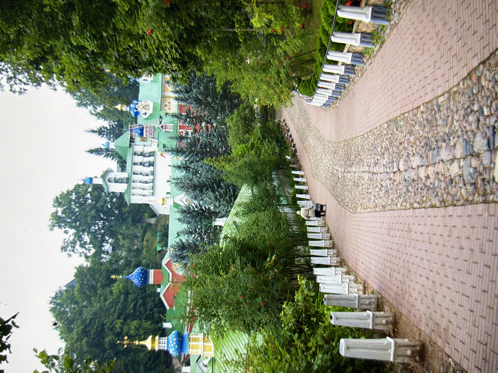 The image depicts a pathway leading towards a complex of buildings with green roofs and colorful domes, characteristic of Orthodox Christian architecture. On the left side of the pathway, there is a series of white decorative pillars. A lone individual, dressed in a light-colored coat and pushing a small trolley, is walking away from the camera along the pathway. The pathway itself is paved with reddish-brown bricks on the right and cobblestones on the left. Lush green trees and foliage surround the pathway and the buildings, suggesting a park-like or garden setting. The sky is overcast, with a pale, diffused light. The architectural style of the buildings, particularly the onion domes, strongly suggests a location within Ukraine, likely a monastery or a significant religious site. Given the distinct architectural style of the buildings, this scene is likely the Kyiv Pechersk Lavra (Monastery of the Caves) in Kyiv, Ukraine, a UNESCO World Heritage site renowned for its extensive complex of churches and caves.