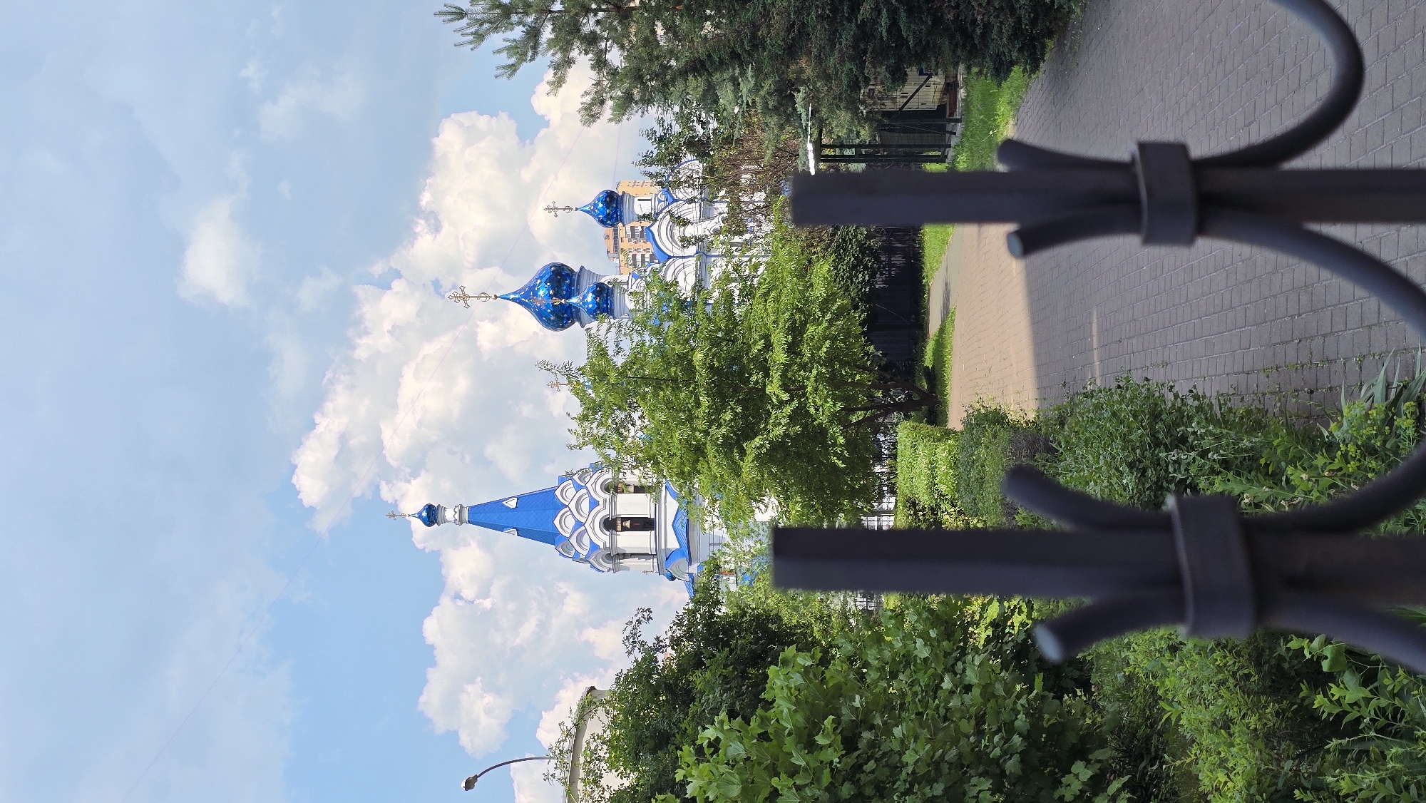 This is a vertical, eye-level, outdoor shot of an Orthodox church in Kyiv, Ukraine, viewed through a dark metal fence in the foreground. The church has a white facade and a blue roof with multiple onion domes topped with crosses. The central dome is a tall spire, while others are rounded. Green trees and bushes are in the foreground and middle ground, with a paved walkway leading towards the church. The sky is blue with scattered white clouds. A street lamp is visible on the left. The scene appears to be during daylight.