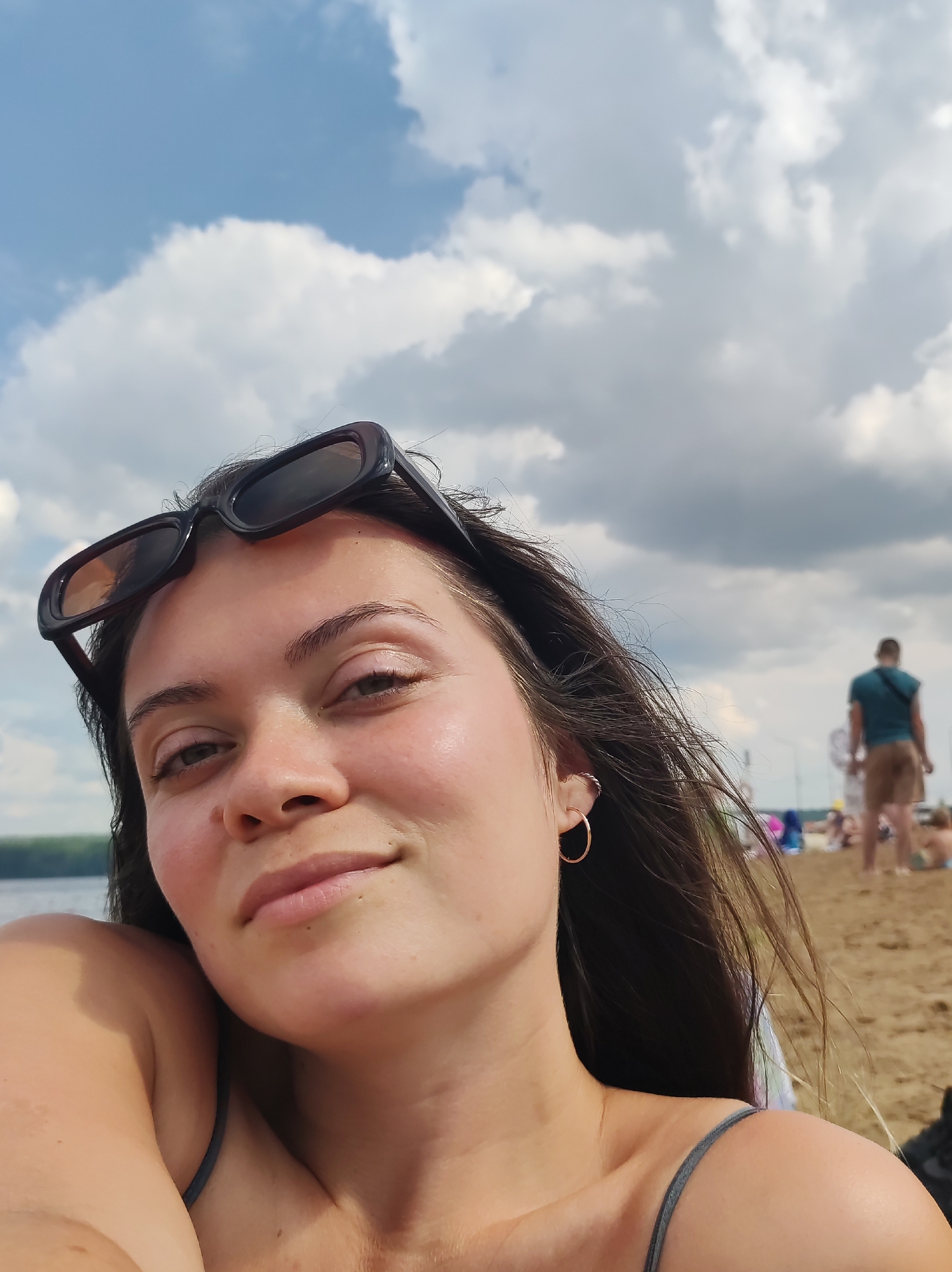 A young woman with dark hair and sunglasses pushed up on her head is taking a selfie on a sandy beach under a cloudy sky. She is wearing a grey spaghetti strap top. In the background, several people are visible in the distance on the beach, some appearing to be sunbathing or walking. A body of water, likely a river or lake, is visible to the left of the frame. The setting is consistent with a beach area in Chaikovsky, Russia.