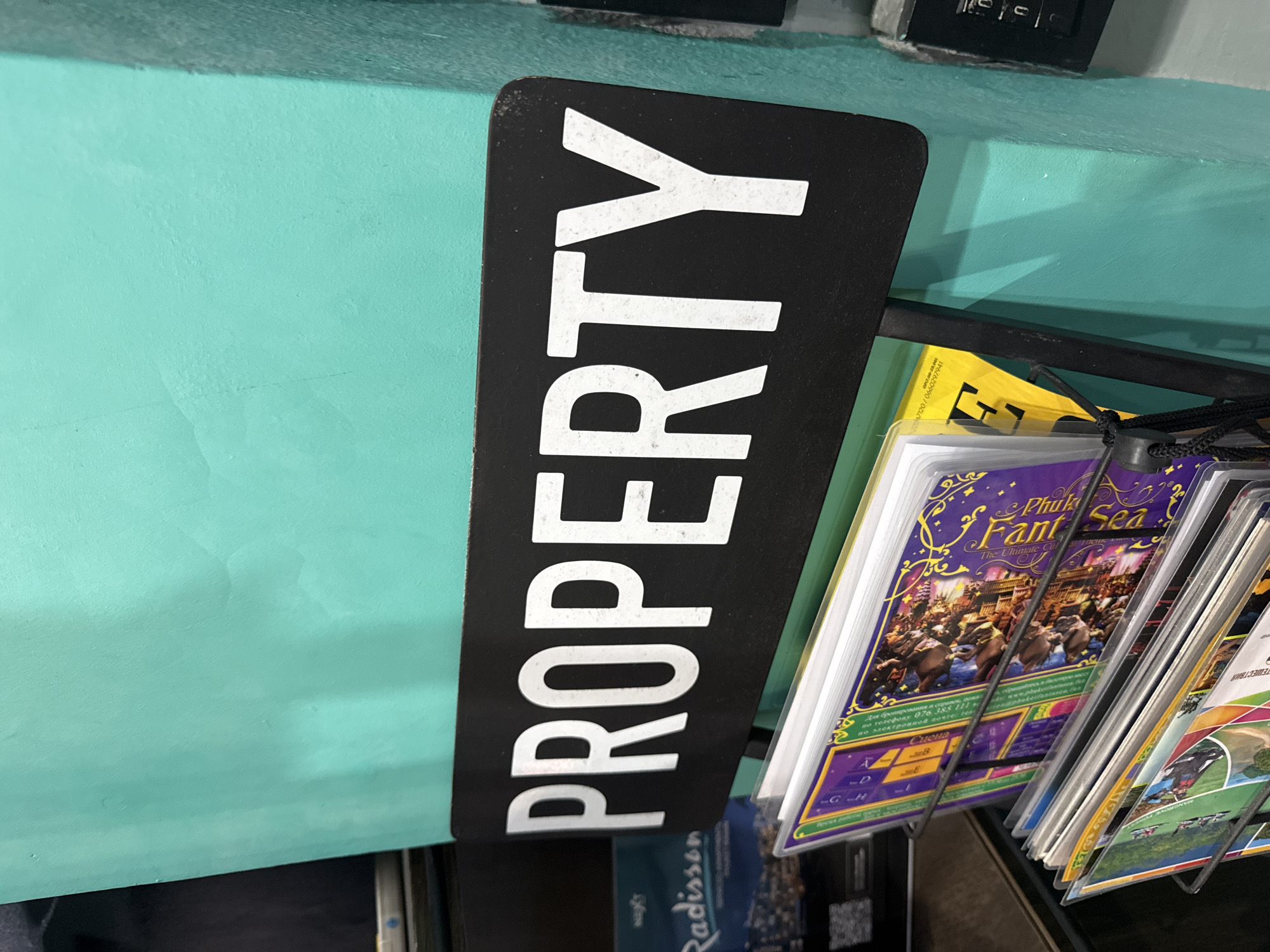 This is a close-up shot of a black sign with white lettering that reads "PROPERTY" in large, capitalized letters, oriented vertically. The sign is attached to a teal-colored wall. To the right of the sign, there is a rack displaying several brochures or pamphlets. One prominent brochure features a vibrant purple and gold design with the text "Phuk Fant Sea" and an illustration of elephants. Other brochures show various scenic images and text in different languages, suggesting a tourist or travel-related context. The overall impression is a display in a hotel lobby or tourist information center. The text on the brochures includes what appears to be booking information in Russian, with a phone number and email address. The background is a plain teal wall, and a dark shelf or counter is visible at the bottom. The lighting suggests an indoor setting, possibly during the day. There is no indication of people or weather in the immediate frame.