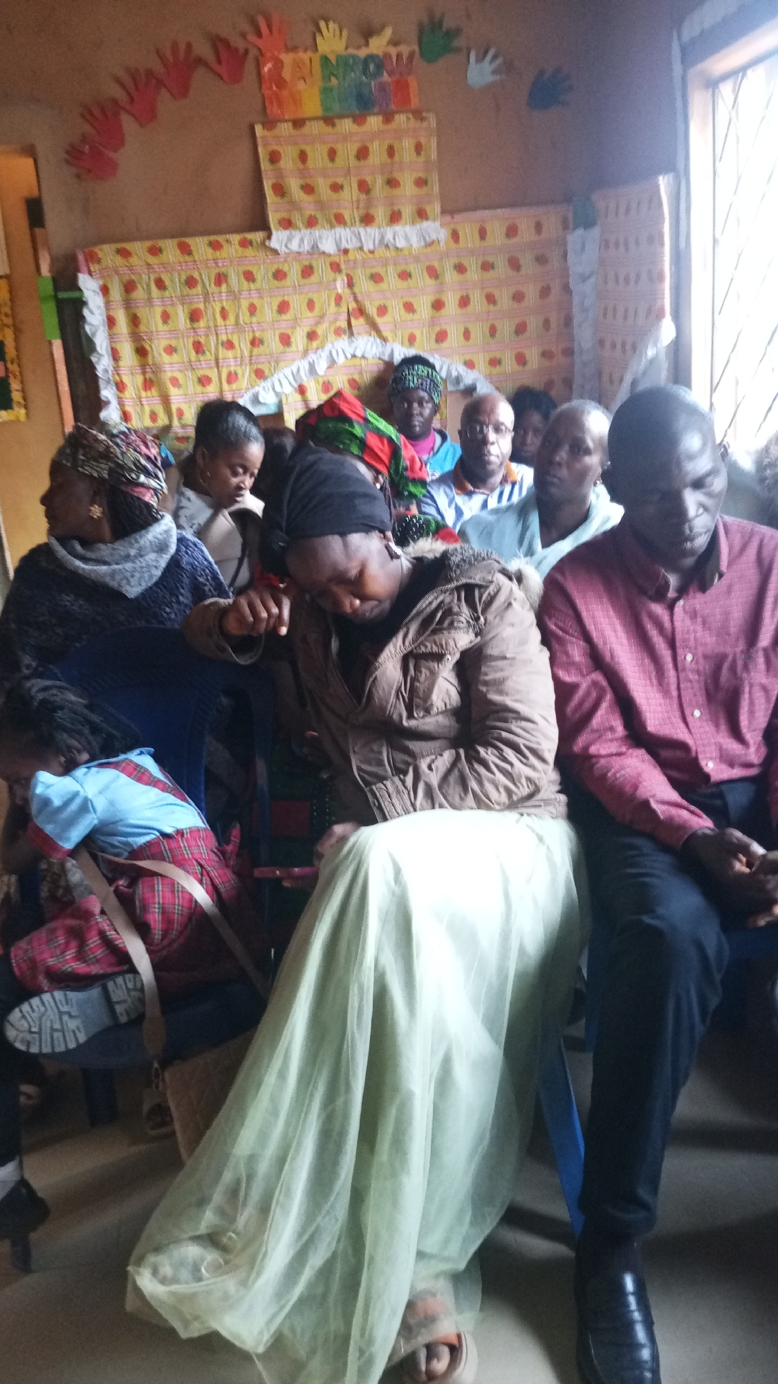 A medium shot shows a group of people sitting in a room, likely a classroom, with decorations on the wall. In the foreground, a woman in a brown jacket and light green skirt is seated, with her head bowed. To her right, a man in a maroon checkered shirt and dark pants is also seated, looking down. To the left of the woman in the brown jacket, a child in a plaid skirt and blue shirt is seated, leaning forward. Other individuals are visible in the background, some of whom appear to be children. The wall behind them is decorated with colorful paper cutouts, including handprints and a sign that reads "RAINBOW". Fabric with a yellow and red strawberry pattern hangs on the wall. A window with bars is visible on the right side of the image. The room appears to be sparsely furnished, with chairs and a bag visible.