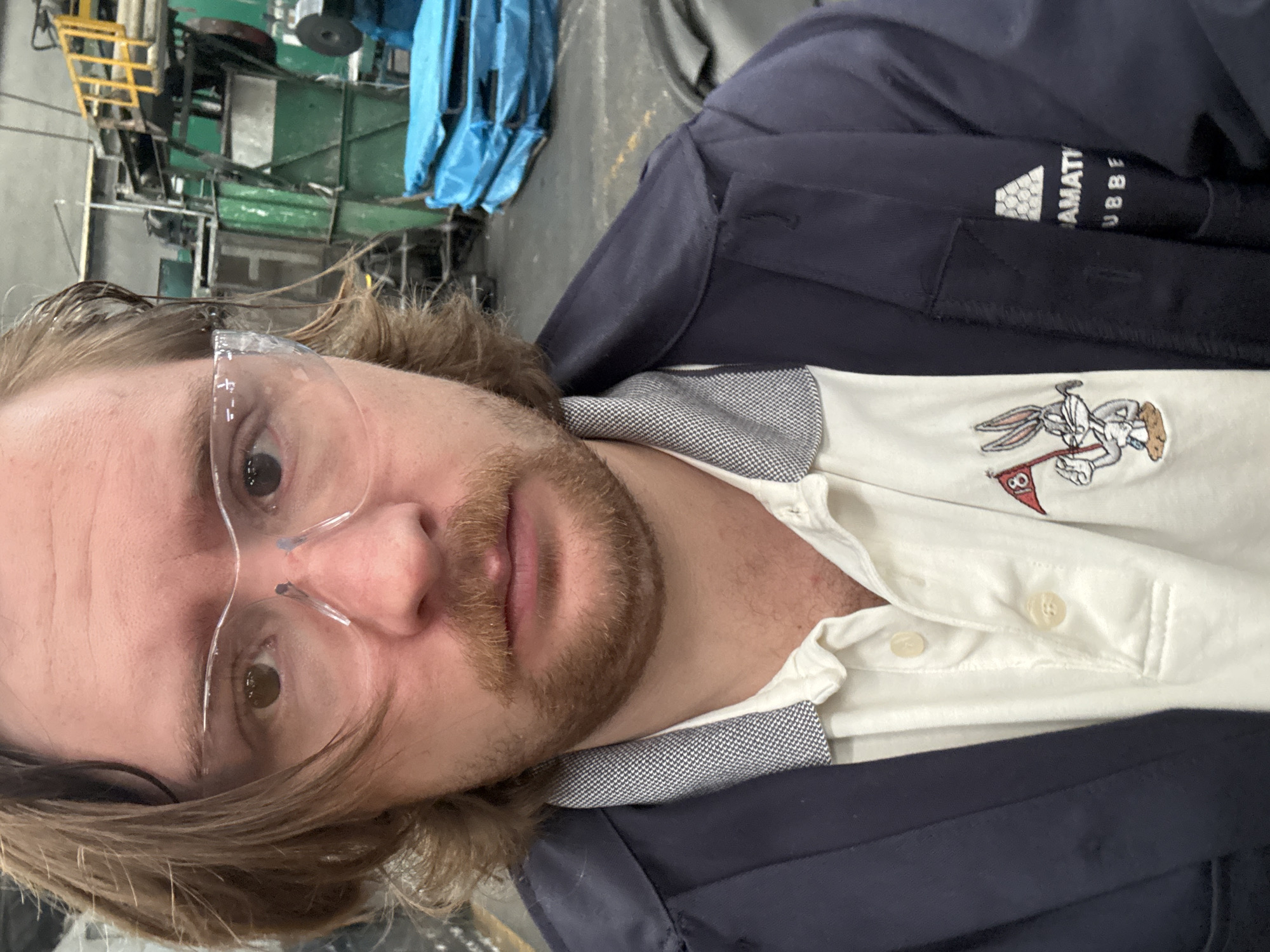 A close-up, rotated selfie shows a man with fair skin, brown hair, and a beard wearing safety glasses. He is wearing a white polo shirt with a Bugs Bunny golf logo and a navy blue jacket. The jacket has "AMATTI RUBBE" on the arm. The background is a blurred industrial setting with green machinery and blue tarps. While the location in Cuautitlán Izcalli, Mexico, is unspecified in the image itself, the information can be considered relevant context.