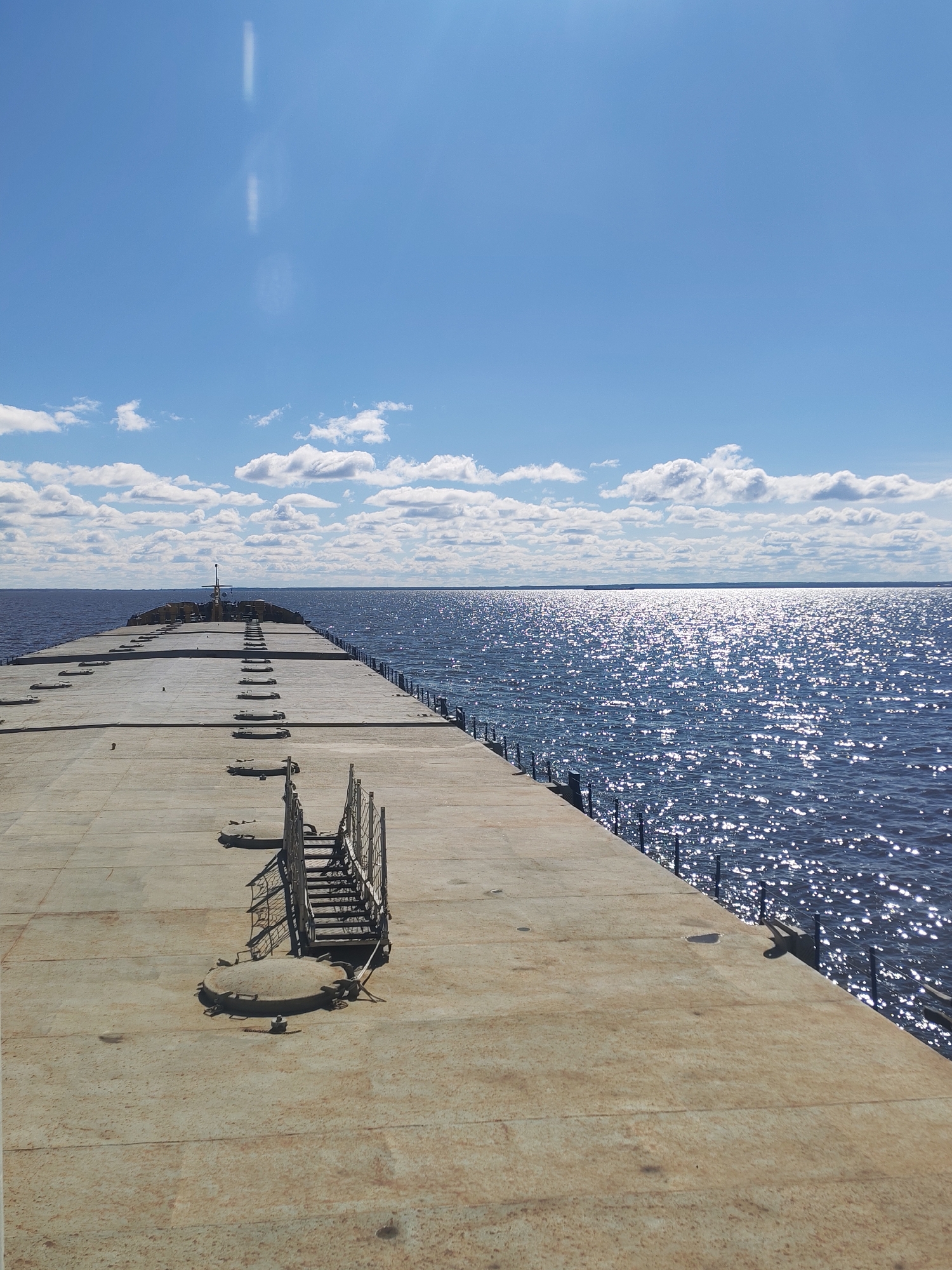 The image depicts a long, concrete pier extending into a body of water, possibly Lake Beloye. The pier is lined with a metal railing on one side and features a series of regularly spaced access points or hatches along its surface. A metal ladder structure is prominently positioned on the pier. The water is sparkling with sunlight, and the sky is partly cloudy. The location is likely Belozersk, Russia, given the characteristics of Lake Beloye and similar structures in the area. There are no people visible in the frame.