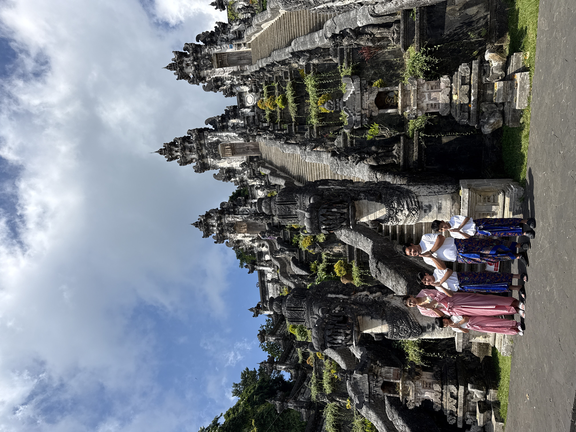 A family of five is posed in front of a large, ornate stone temple complex in Abang, Indonesia. The family consists of two adults and three children, all dressed in traditional Balinese attire. The adult male wears a white shirt and a blue and gold sarong, and the adult female wears a pink wrap dress and a sarong. The children also wear white shirts and sarongs in coordinating colors. All five individuals have their hands pressed together in a prayer-like gesture.
The temple complex is characterized by intricate stone carvings, multi-tiered roofs, and imposing staircases adorned with dragon motifs. Lush green vegetation is visible throughout the structure, with some plants cascading down the stone walls. The sky above is a mix of blue with dramatic white clouds. The ground in front of the temple is paved with dark stone. The overall scene captures a moment of cultural and spiritual significance, with the family participating in a traditional custom at a historically rich location.