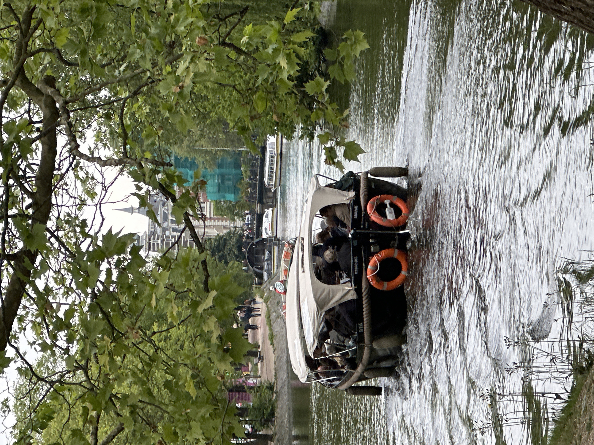 A boat filled with people is traveling on a river in Amsterdam, Netherlands. The boat has a white canopy and orange life preservers attached to the back. A tree with green leaves is visible in the foreground on the left. Buildings and other boats can be seen in the background. People are walking along the riverbank on the left side of the image. The water is rippling from the boat's movement.
