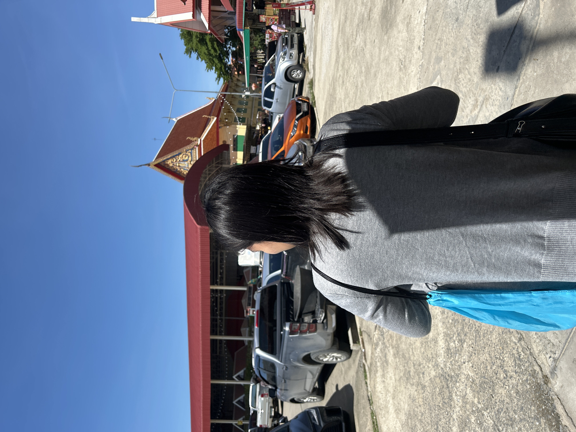 A woman with long, dark hair is seen from behind, walking outdoors in what appears to be a parking lot. She is wearing a grey sweater and carrying a bright blue bag slung over her left shoulder, along with a black shoulder bag. In the background, several vehicles, including pickup trucks and cars, are parked. Beyond the vehicles, there is a large building with a red roof and a more ornate structure with a multi-tiered, curved roof that is characteristic of Thai temples. The sky is clear and blue. This scene is located in the city of Thai Ko Subdistrict, Thailand.