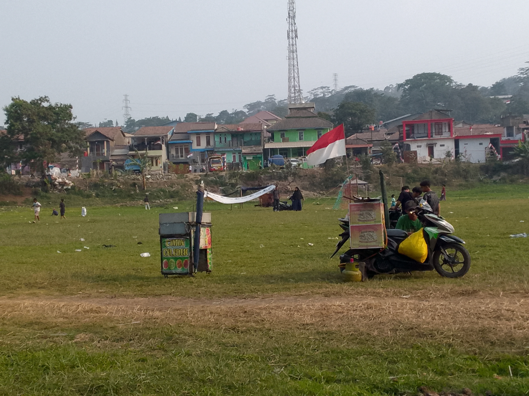 In Ciburuy, Indonesia, a scene unfolds with a backdrop of a residential area characterized by a mix of single-story and multi-story buildings, some painted in bright colors like green and red, others in more muted tones. A tall telecommunications tower dominates the skyline in the distance, alongside a hazy, overcast sky. In the foreground, a large grassy field with scattered litter and some dry patches suggests a park or open space.
Several people are present in the image. Near a white tent-like structure, a group of individuals are gathered, some appearing to interact. Further into the field, a few individuals are seen standing and walking.
Dominating the right side of the frame is a motorcycle with a large yellow bag strapped to its seat and two stacked boxes on the rear rack. The boxes are labeled with text that is difficult to read clearly. A green gas cylinder is positioned near the motorcycle. To the left of the motorcycle, a mobile food cart with a green exterior and a sign displaying "CIMIN CELEDEK" and circular icons is visible. Several people are near or interacting with this cart and the motorcycle.
The overall impression is a casual gathering or market scene in a semi-urban Indonesian setting, with elements of daily life and commerce on display in an open, grassy area.