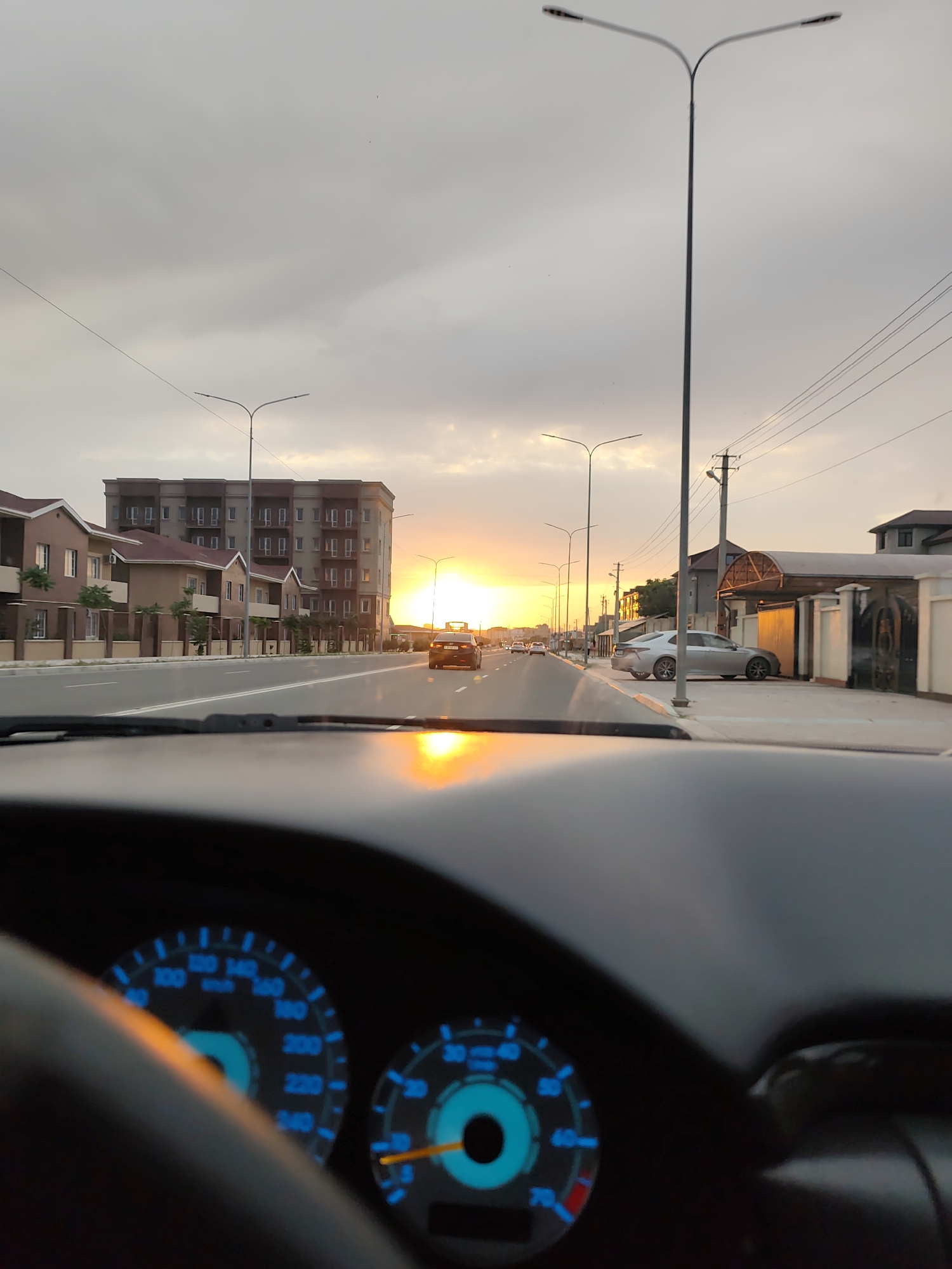 The image captures a view from inside a car looking out onto a street in Ömirzaq, Kazakhstan, during sunset. The sky is filled with a gradient of colors from soft grays to bright oranges, indicating the sun is low on the horizon. Scattered clouds add texture to the sky.
The scene depicts a residential and commercial area. On the left side of the street, there are modern, multi-story apartment buildings and smaller houses with red-tiled roofs. On the right side, there are more houses and some structures with covered parking areas, suggesting a suburban or developing urban environment. Streetlights line both sides of the road, and power lines are visible against the sky.
Several cars are visible on the road, indicating normal traffic flow. One car is driving away from the camera in the distance, and another is parked on the right side of the street. The dashboard of the car from which the photo is taken is partially visible in the foreground, with illuminated gauges showing speed and RPMs. The reflection of the sunset is also prominent on the dashboard and windshield, adding warmth and color to the interior view.
The time of day is clearly sunset, with the warm glow of the sun casting long shadows and illuminating the surroundings. The weather appears to be clear, with the sunset being the most striking atmospheric element. The location cues, such as the architectural style of the buildings and the presence of streetlights and power lines, are consistent with a developed area in Kazakhstan. No people are clearly discernible. The overall mood of the image is peaceful and scenic, highlighting the beauty of the sunset over a contemporary landscape. There is no visible text in the image.