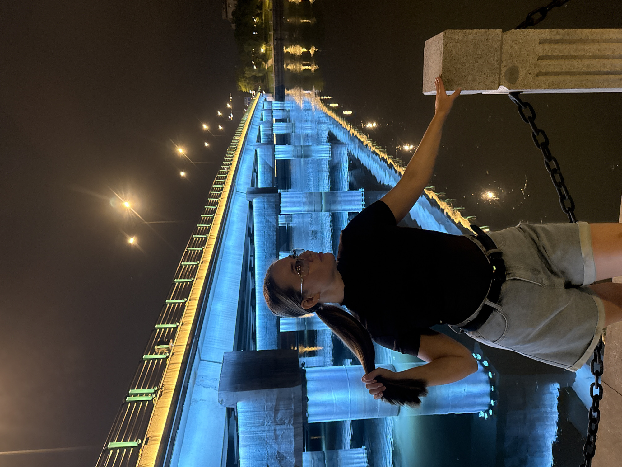 A woman stands on a walkway next to a body of water at night. The walkway has a stone pillar and a chain railing. Behind her, a bridge is illuminated with blue and yellow lights, and its reflection is visible in the water. The woman is wearing a black t-shirt, denim shorts, and glasses. She is holding her ponytail with her left hand and resting her right arm on the stone pillar. The scene appears to be in Yiwu, China, possibly near the Yiwu River.