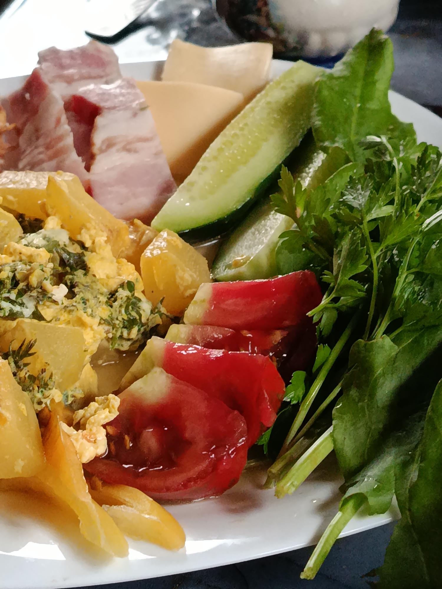 A close-up, overhead view of a white plate holds a meal consisting of several distinct components. To the left, there are slices of bacon, a light yellow cheese, and several pieces of cooked yellow potato. Next to the potatoes is a portion of scrambled eggs mixed with green herbs. In the center and right of the plate are slices of red tomato, cucumber slices, and a generous serving of fresh green parsley. The lighting is somewhat dim, suggesting an indoor setting, possibly a table. The specific location is Unknown, Unknown.