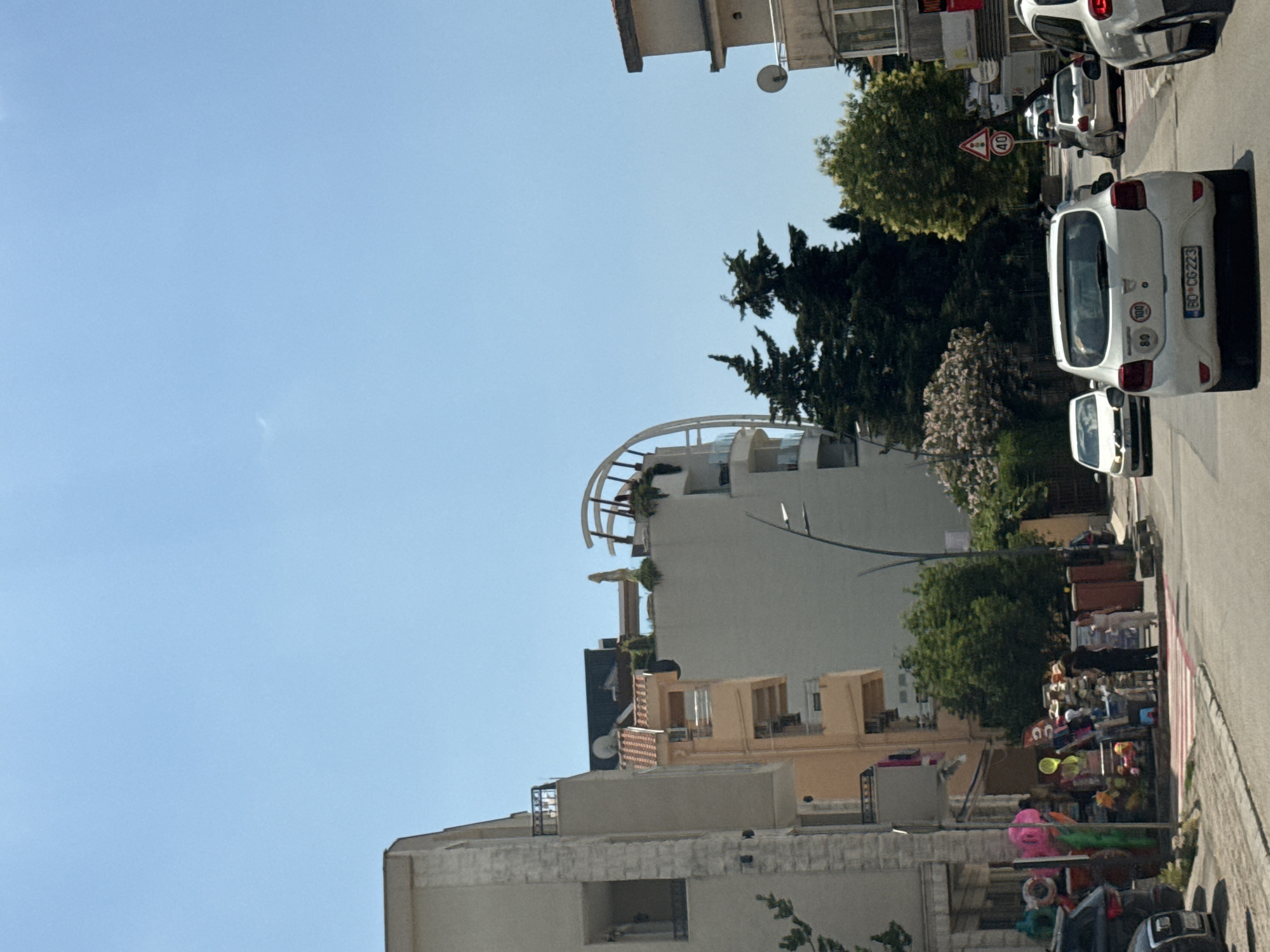 The image shows a street scene in Budva, Montenegro, on a clear, sunny day. The foreground features a road with several cars. A white Dacia Sandero is prominent in the center, with its license plate visible. To the left, a dark-colored SUV is parked. In the distance, other vehicles are on the road, and there are signs, including a speed limit sign indicating "40".
The left side of the street is lined with buildings, one of which appears to be a multi-story apartment building with balconies. A shop selling beach items and souvenirs is visible at street level, with various colorful items displayed. Two people are seen near the shop.
On the right side of the street, more buildings are visible, including a light-colored apartment building with a unique curved architectural feature on its upper floors. Trees, including some tall evergreen trees and leafy deciduous trees, are scattered throughout the scene, providing a contrast to the urban architecture. The sky is a bright, clear blue. The overall impression is a bustling street in a coastal town during daytime.