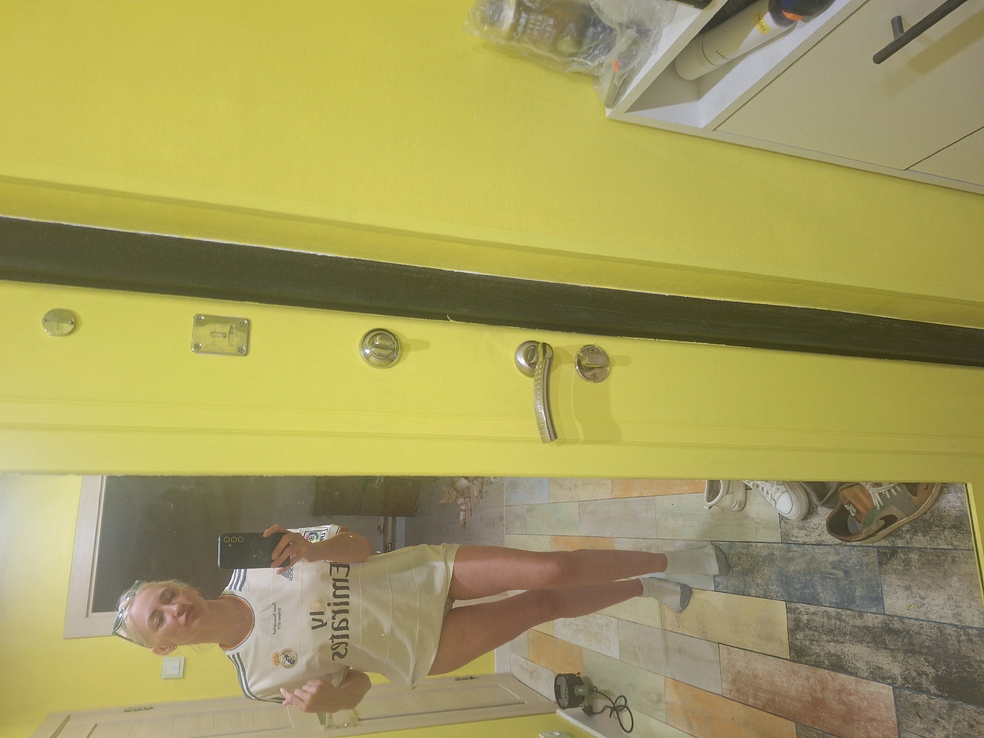 An adult Caucasian female is taking a selfie in a mirror. She is wearing a white t-shirt that appears to be a sports jersey and white socks. She has blonde hair and is wearing sunglasses on top of her head. The background shows a bright yellow wall, a mirror, a door with a silver handle and lock, a white shelf unit with items on it, and a floor with multi-colored wooden planks. A ginger cat is visible in the reflection of the mirror, lying on the floor. The scene appears to be an indoor domestic setting, potentially an apartment or house. The provided location of Kaluga, Russia, is not directly verifiable from the visual information in the image.