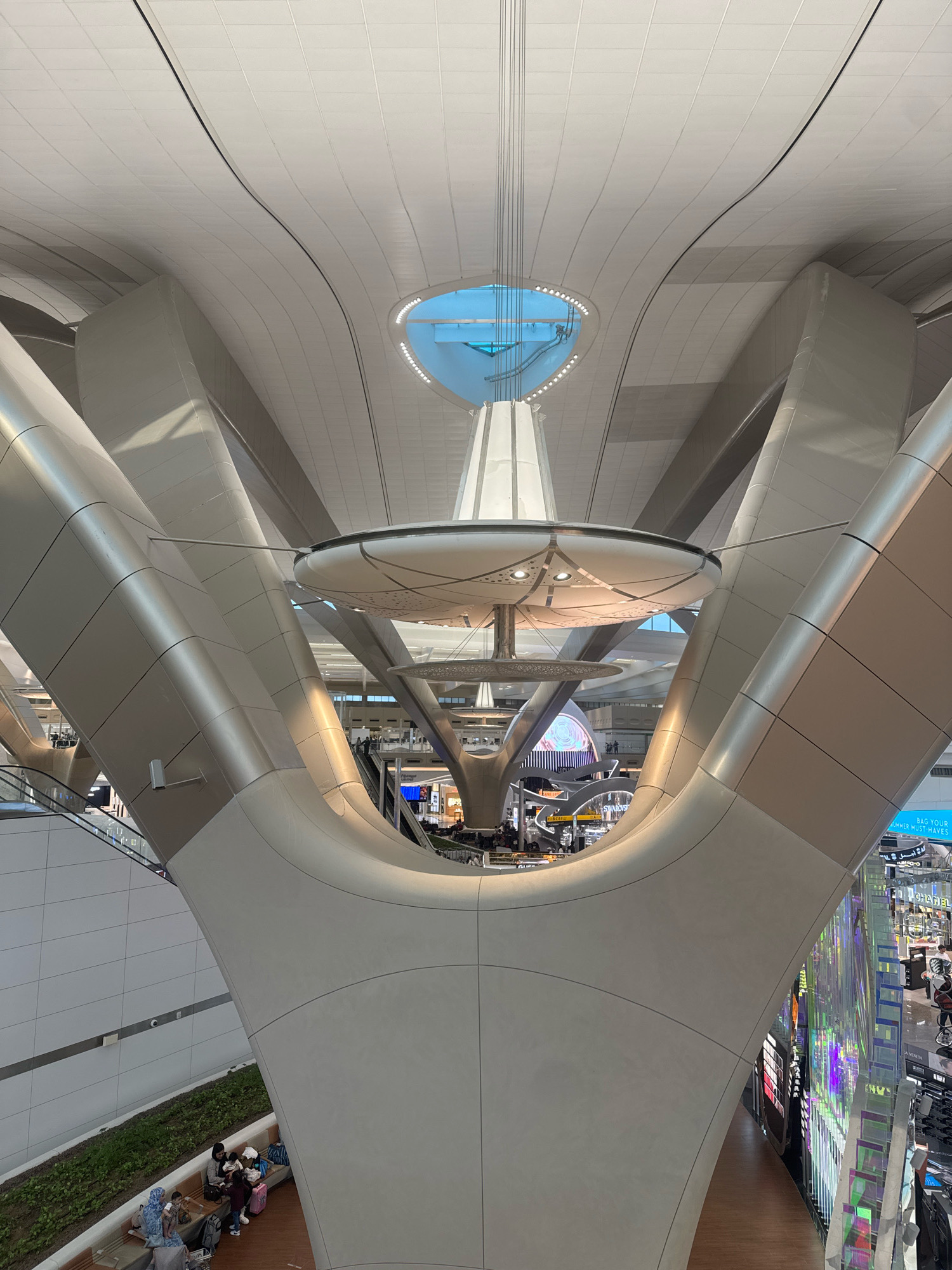This is an interior, eye-level, vertical shot of the Hamad International Airport in Al Matar, United Arab Emirates. The airport features a modern, futuristic architectural design with a white, curved ceiling and large, beige, sculpted support columns. Suspended from the ceiling are circular, multi-tiered light fixtures. In the background, escalators are visible, along with various shops, signs, and people. On a lower level, a group of people are seated on benches surrounded by greenery. The lighting is bright, reflecting off the polished surfaces.