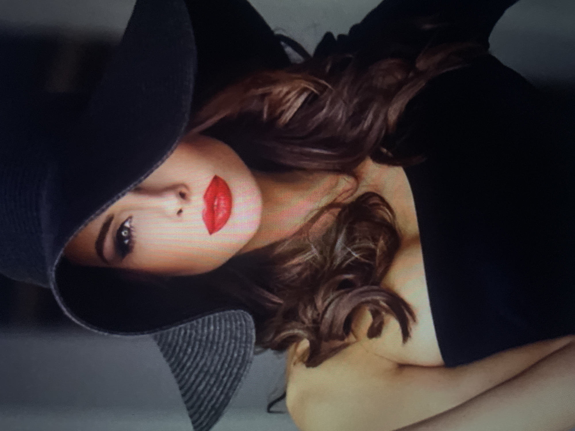The image is a close-up portrait of a woman wearing a wide-brimmed black hat and a black off-the-shoulder top. She has dark, wavy hair that falls around her shoulders. Her makeup is dramatic, with bold eyeliner and bright red lipstick. The background is a muted gray, creating a focus on the woman. There is no identifiable information in the image that places it in the city of Kołobrzeg, Poland.