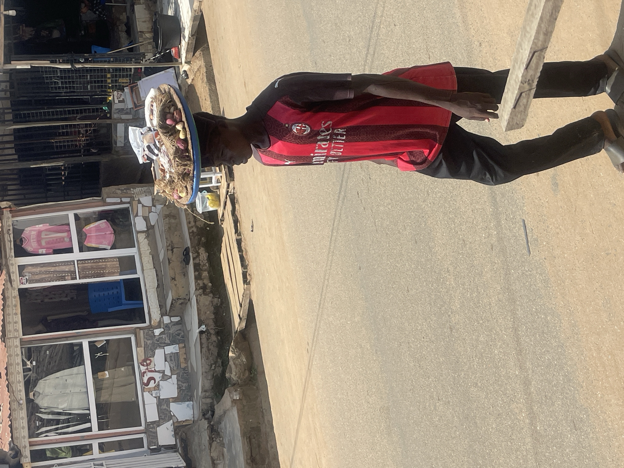 A young man, likely in his late teens or early twenties, walks along a paved street in Abuja, Nigeria. He is wearing a red and black striped soccer jersey and black trousers. Balanced on his head is a large, circular tray filled with various food items, including what appear to be yams, onions, and other produce, some of which are wrapped in netting or plastic. In the background, a small shop with a metal grate enclosure displays clothing in its windows. The ground is a mix of asphalt and dirt, with some visible debris. The overall lighting suggests it is daytime, with shadows indicating the sun is present.