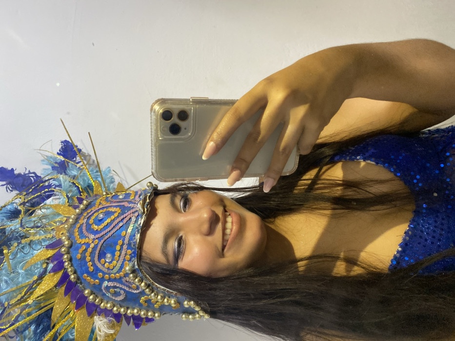 A smiling young woman takes a selfie. She wears a blue sequined top and an elaborate feathered headdress with gold accents and bead embellishments. Her dark hair falls across her chest. She holds a smartphone with a clear case in her right hand. The background is a plain white wall.