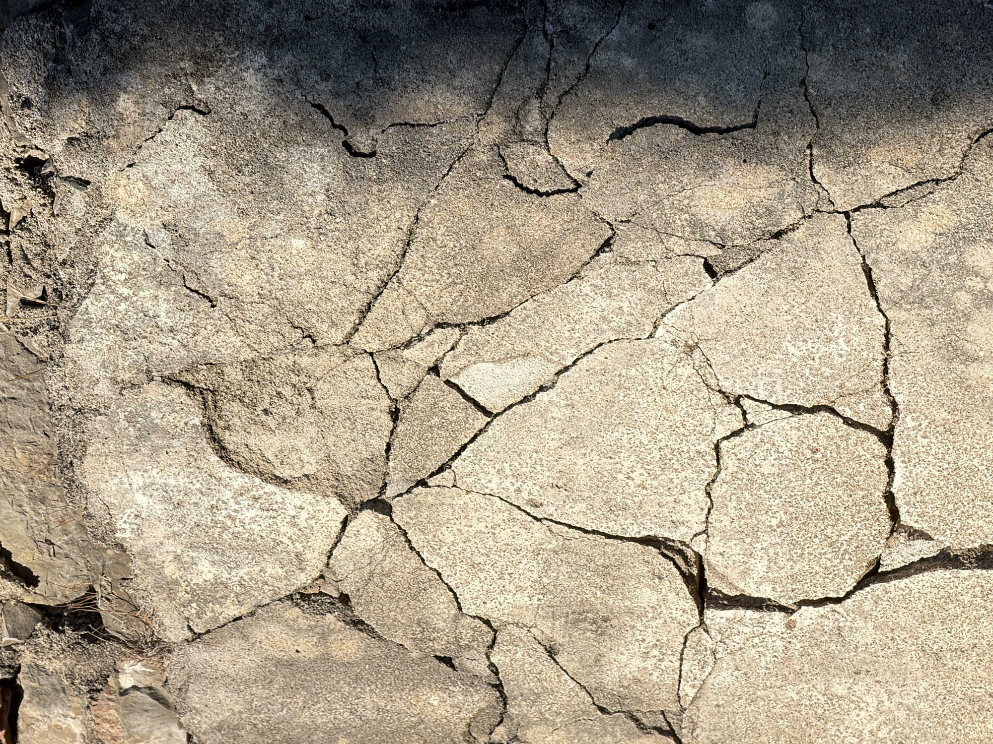 The image shows a close-up view of a cracked, weathered concrete surface. The concrete appears to be part of an outdoor structure, possibly a wall or pavement, given the presence of small stones and dried vegetation at the top edge of the frame. The cracks are numerous and irregular, dividing the surface into various polygonal fragments. Some areas of the concrete are lighter and appear rougher, while others are darker and smoother, with visible shadows cast across the surface, suggesting an overhead light source. The overall impression is one of age and decay. The location is identified as Androvići, Montenegro.