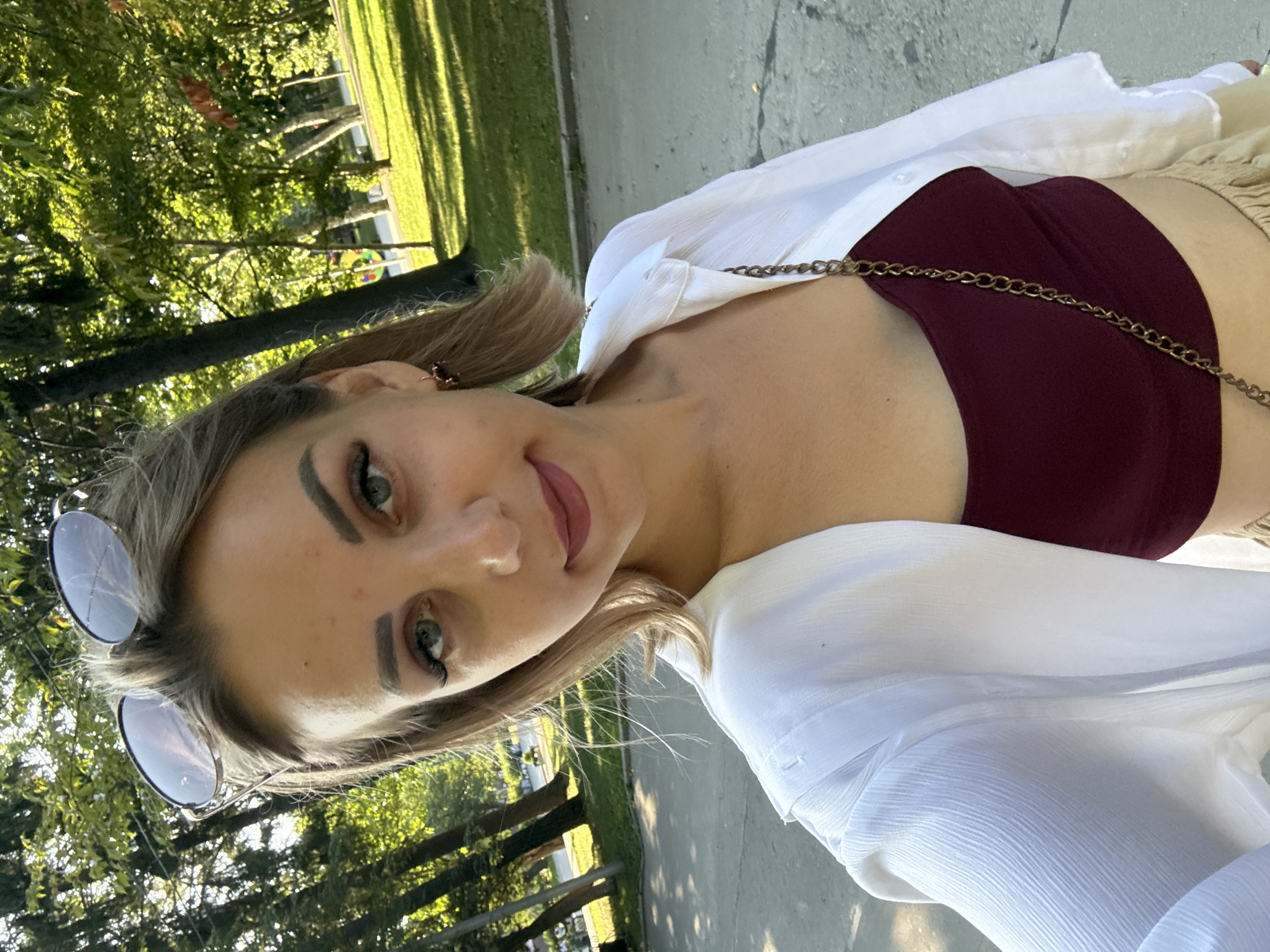 The image shows a young Caucasian woman with blonde hair and green eyes posing for a selfie outdoors. She is wearing a white open button-down shirt over a burgundy crop top and a light-colored skirt. Sunglasses are perched on top of her head. The background features a park with trees and a paved walkway, suggesting a public outdoor setting. The lighting indicates it is daytime, with sunlight filtering through the leaves of the trees, casting shadows on the ground. The woman's facial expression is neutral. The specific location in Yuzhno-Sakhalinsk, Russia, cannot be determined from the visual information alone, as there are no identifying landmarks or textual elements present in the image.