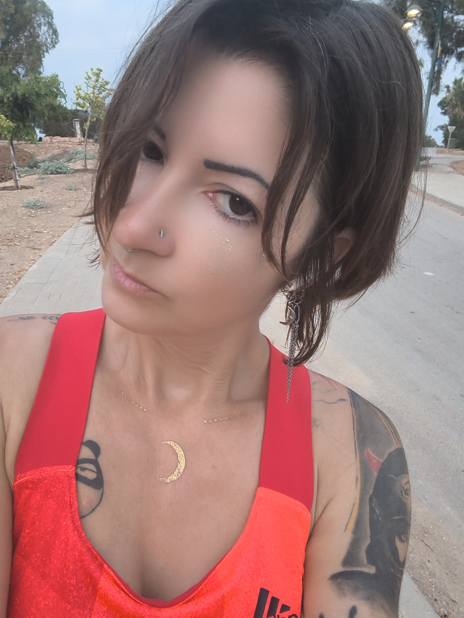 A medium shot captures a woman from the chest up, facing slightly towards the camera. She has dark brown, chin-length hair that is styled loosely, with some strands falling across her face. She is wearing a bright red, sleeveless top. A delicate gold necklace with small circular charms is visible around her neck. She has a tattoo on her chest to the left of the necklace, appearing to be a stylized panda face. To the right of the necklace, a gold crescent moon design is visible on her skin. She has a nose ring and an earring on her left ear, which appears to be a silver chain with a moon charm. Her right arm, partially visible, has extensive tattoos. She is outdoors, with a paved pathway behind her and a dirt and gravel area to her left. In the background, there are trees, lampposts, and a glimpse of a body of water, suggesting a park or recreational area. The location is identified as Lachish Regional Council, Israel.