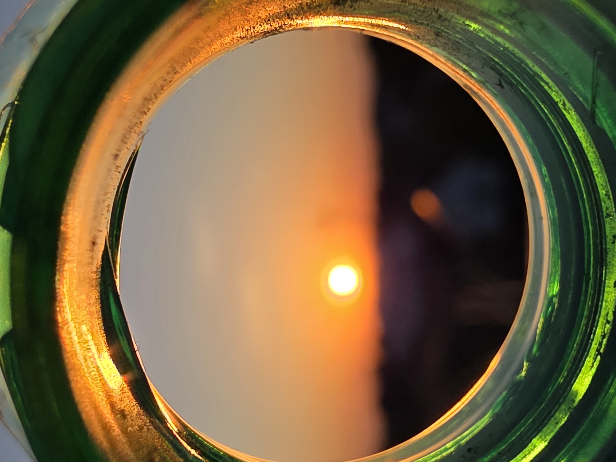 A close-up shot depicts the sun viewed through the opening of a green glass object, possibly a bottle. The opening of the glass object creates a circular frame. The setting sun is centrally located within the frame, emitting an orange glow. The environment around the sun appears blurred with color gradient on both sides, likely the sky on one side and a dark mass on the other. The green glass object framing the sun shows various degrees of transparency and color gradients depending on the angle of the light. The photo gives an artistic and distorted view of the natural sunlight scene. The location is not discernible from the image.