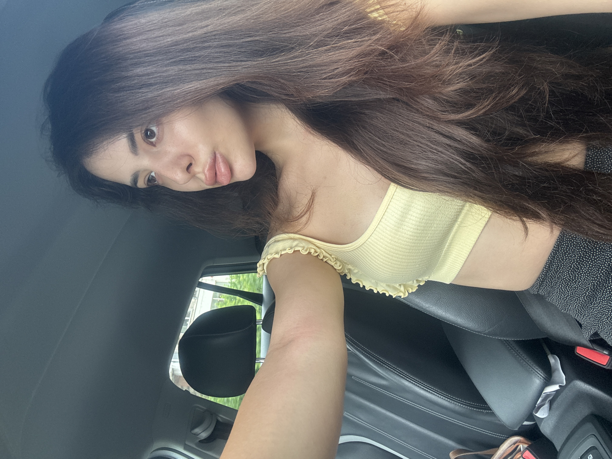 An informal portrait captures a young woman with long, voluminous dark brown hair, positioned inside a car, likely in Plovdiv, Bulgaria. The image is taken at an angle, possibly a selfie. She looks directly at the camera with a subtle, somewhat pouting expression. Her long, dark hair cascades around her, partially covering her face and shoulder. She is wearing a light yellow, ribbed crop top with delicate ruffled trim along the shoulder straps, and a dark, polka-dotted skirt or shorts. Her arm is visible in the foreground, extending towards the camera.
The setting is a car interior, featuring dark leather seats, a headrest, and a visible red and black seatbelt buckle. The lighting is bright, suggesting it is daytime. Through the car window, glimpses of a bright, possibly green, outdoor environment can be seen. There is no visible text in the image.