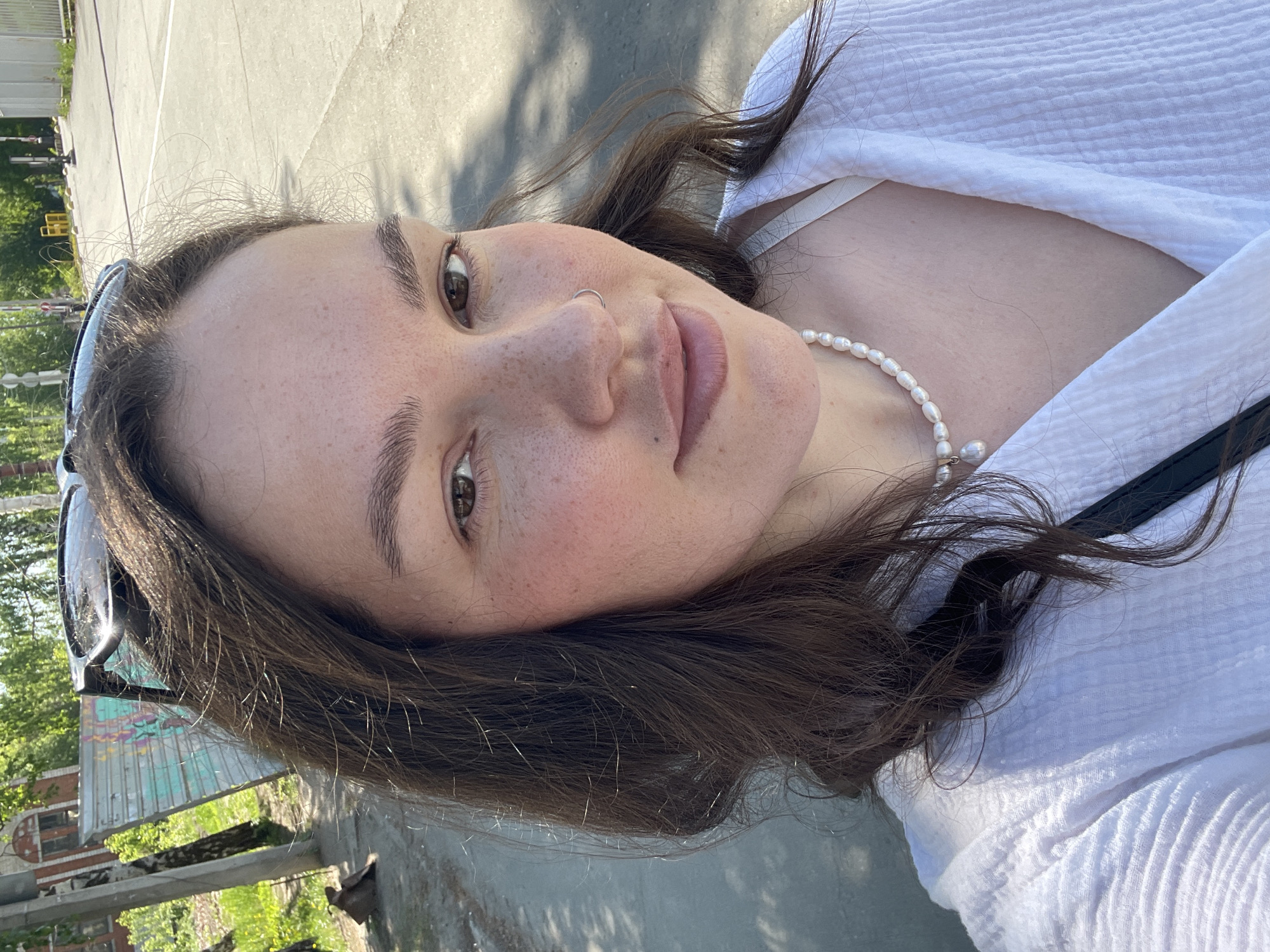 A selfie of a fair-skinned female with brown hair and eyes, wearing a white blouse and a pearl necklace. She has a nose ring on her left nostril. Sunglasses are atop her head. The background is blurred, but suggests an urban setting. The image was taken outdoors, presumably in Izhevsk, Russia, judging by the surrounding architecture which looks to be a small building with graffiti and a bridge on the side. The ground is concrete or asphalt with some shadow, implying a sunny day.