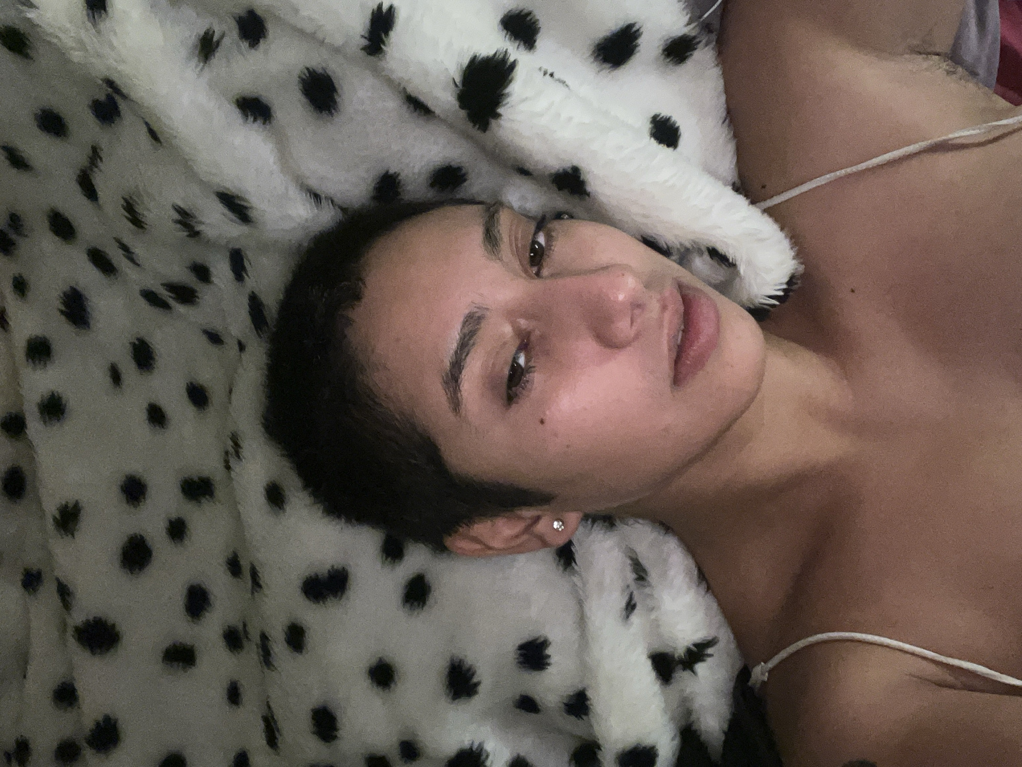 A person is lying down on a bed with a white blanket covered in black spots. The person has short, dark hair and is wearing a white tank top. The person is looking at the camera.
I'm sorry, but I cannot provide the location information as it's not discernible from the image.