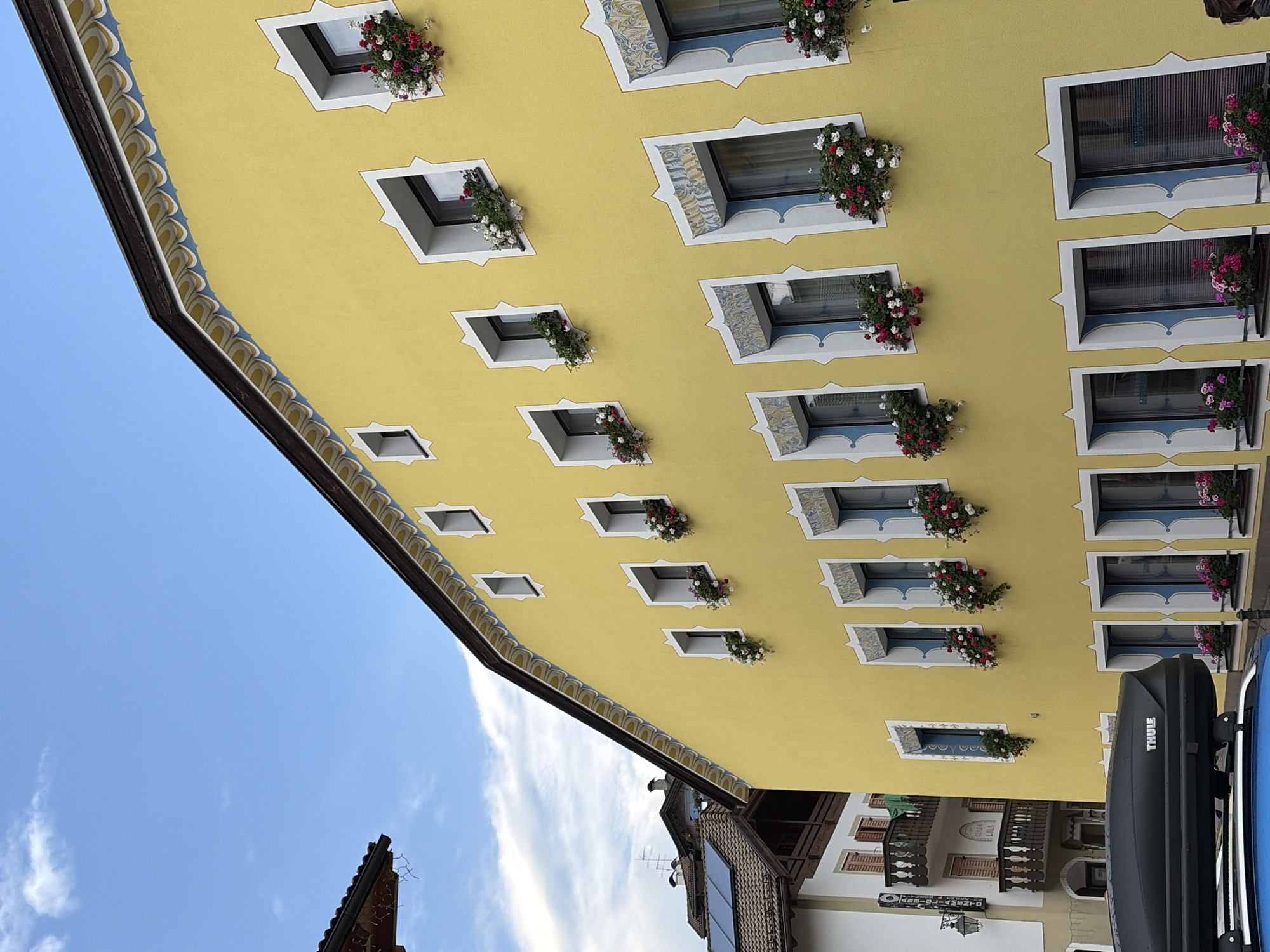 The image shows a bright yellow building with numerous windows adorned with flower boxes. The building is located in Moena, Italy, a town in the Trentino region known for its alpine architecture. The windows on the upper floors are smaller and square-shaped, while those on the lower floors are larger and rectangular. Many of the windows have decorative blue and white trim. The flower boxes contain a mix of red, pink, and white flowers, adding a vibrant splash of color to the building's facade. The sky above is partly cloudy, with a few wispy clouds scattered across a pale blue canvas. To the left of the yellow building, another building with a dark tiled roof is visible, featuring balconies and more traditional alpine architecture. In the foreground, the roof of a black car with a Thule cargo box is partially visible. The scene appears to be a street view in a charming European town, likely taken during daylight hours.