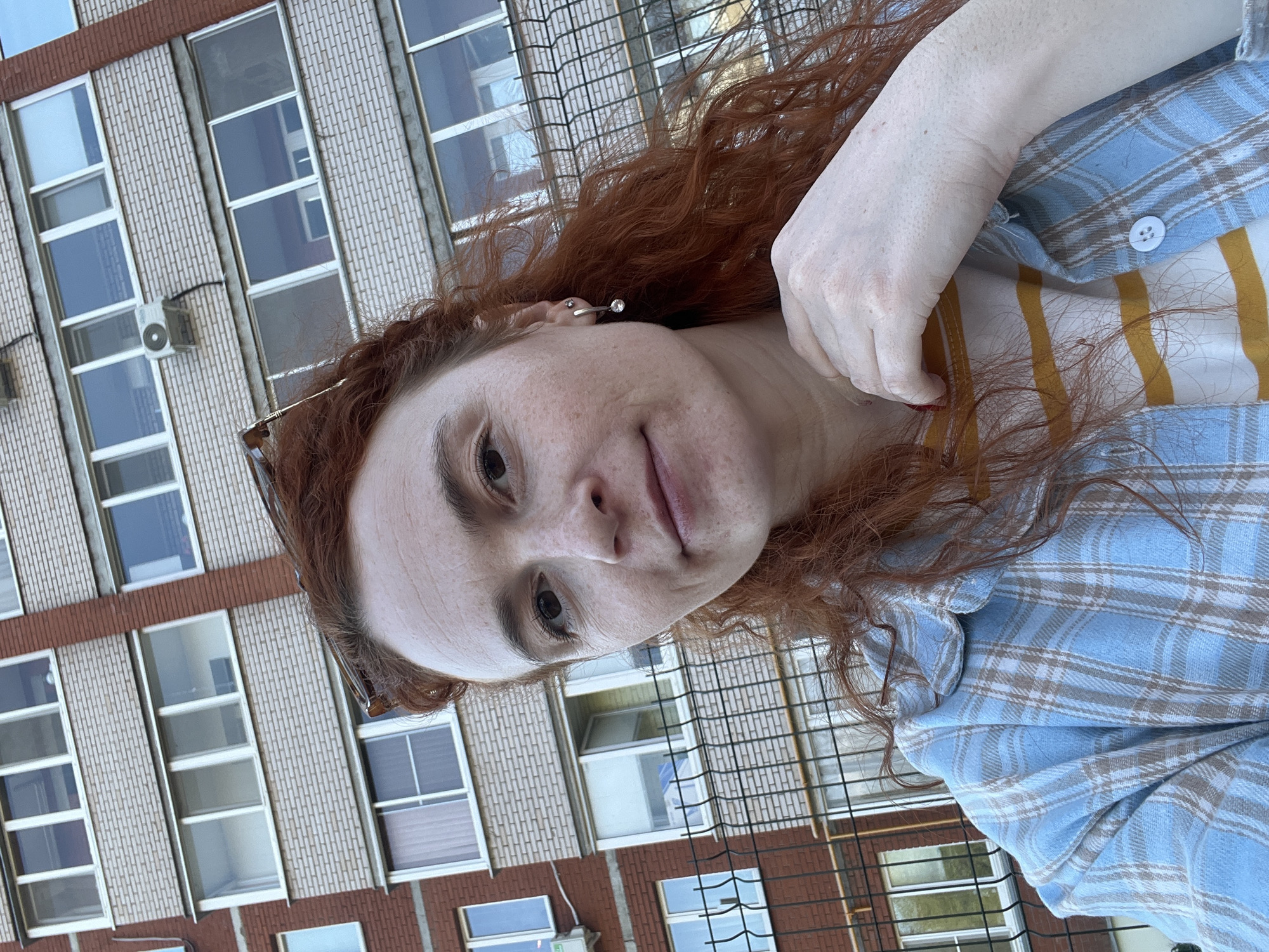 A woman with auburn, curly hair takes a selfie outdoors. She is wearing a yellow and white striped shirt with a light blue and tan plaid button-down over it. She is wearing glasses pushed back on her hair, and a single earring. The background is a brick building with rows of windows, and a fence. The building has the general aesthetic of structures in Chaikovsky, Russia.