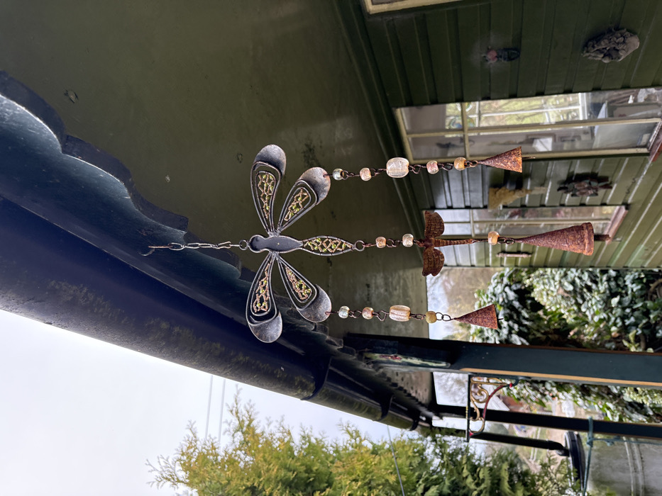 A weathered metal dragonfly wind chime hangs suspended from the dark green eaves of a building in Stiperstones, United Kingdom. The central component is a large, grey-silver metallic dragonfly featuring intricate green-gold patterns within its wings and body. From this, three strands descend, each comprising clear and amber-colored beads, a smaller rustic metal dragonfly, and terminating in a rusty, conical metal bell.
The wind chime is attached via a hook to the underside of a dark green roof overhang, which displays a scalloped edge and a dark, weathered gutter above. The building itself is a dark green wooden structure with vertical siding and multiple windows, reflecting an overcast sky and surrounding vegetation. Several small, rustic metal decorations, including a leaf or bird motif and a pinecone shape, are affixed to the exterior wall of the building. To the left, a dark vertical support column with an attached decorative metal bracket is visible. The background consists of dense green foliage and trees under an overcast sky, with faint utility lines discernible in the upper left. No individuals are present in the scene.