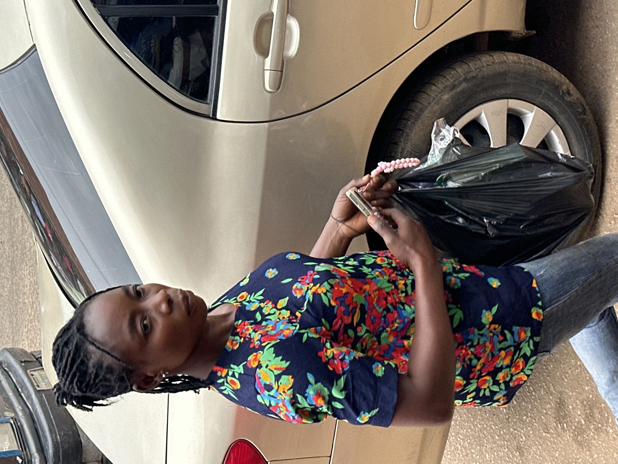 A woman with braided hair stands next to a gold sedan in Bukuru, Nigeria. She is wearing a dark blue shirt with a floral pattern and blue jeans. In her hands, she holds a phone and a black plastic bag. The woman is looking away from the camera. The car's rear passenger door is open, and the tire is visible. The ground is a dusty road.