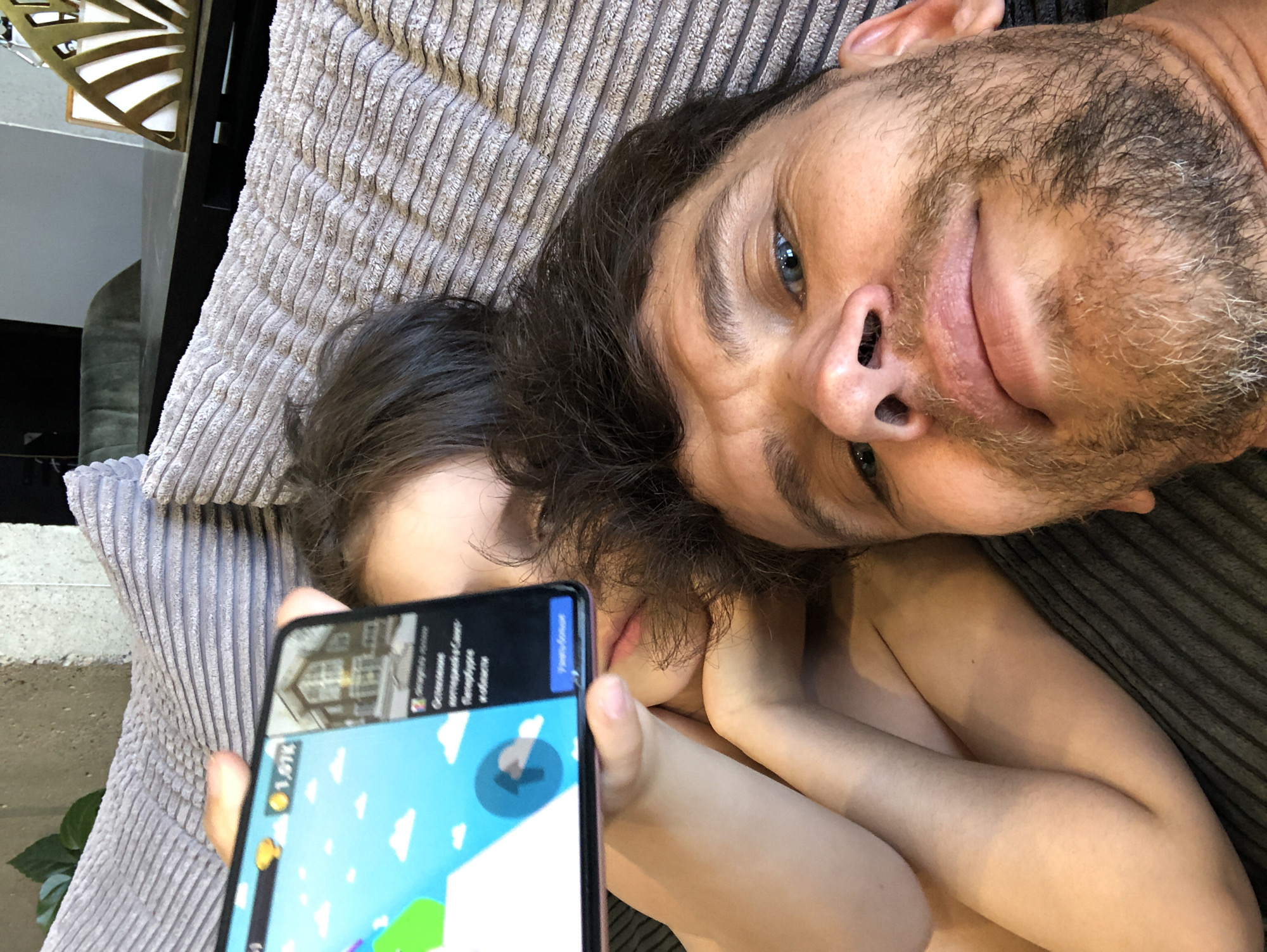 An adult male and a child or young person are captured in a close-up selfie-style image, both lying on a grey, ribbed textile surface, possibly a sofa or bed. The adult male, in the foreground, has blue eyes, dark hair, and a short, mixed brown and grey beard. His face shows mild wrinkles around the eyes, and he is looking directly towards the camera. A child or young person is partially visible behind a horizontally held, dark-framed smartphone, with dark hair, one eye, and lips discernible. The child's hand is holding the device.
The smartphone screen displays two distinct applications. The left side shows a mobile game interface with a light blue sky, white clouds, green and purple game elements, a yellow trophy icon, and the number "1.97k". A blue circular icon with a white upward arrow is also present. The right side of the screen displays an advertisement featuring a house image, underneath which Russian text is visible. This text includes "Объявления" (Advertisements), "выберите город" (choose city), and "Санкт-Петербург и область" (Saint Petersburg and region), followed by "Узнать больше" (Learn more).
In the background, to the upper right, dark furniture, a golden fan-shaped decorative item, and a dark green upholstered chair are partially visible. To the upper left, a green plant and a rough grey wall are present. The setting is an indoor residential environment, specifically located in Saint Petersburg, Russia, as indicated by the text on the phone screen.