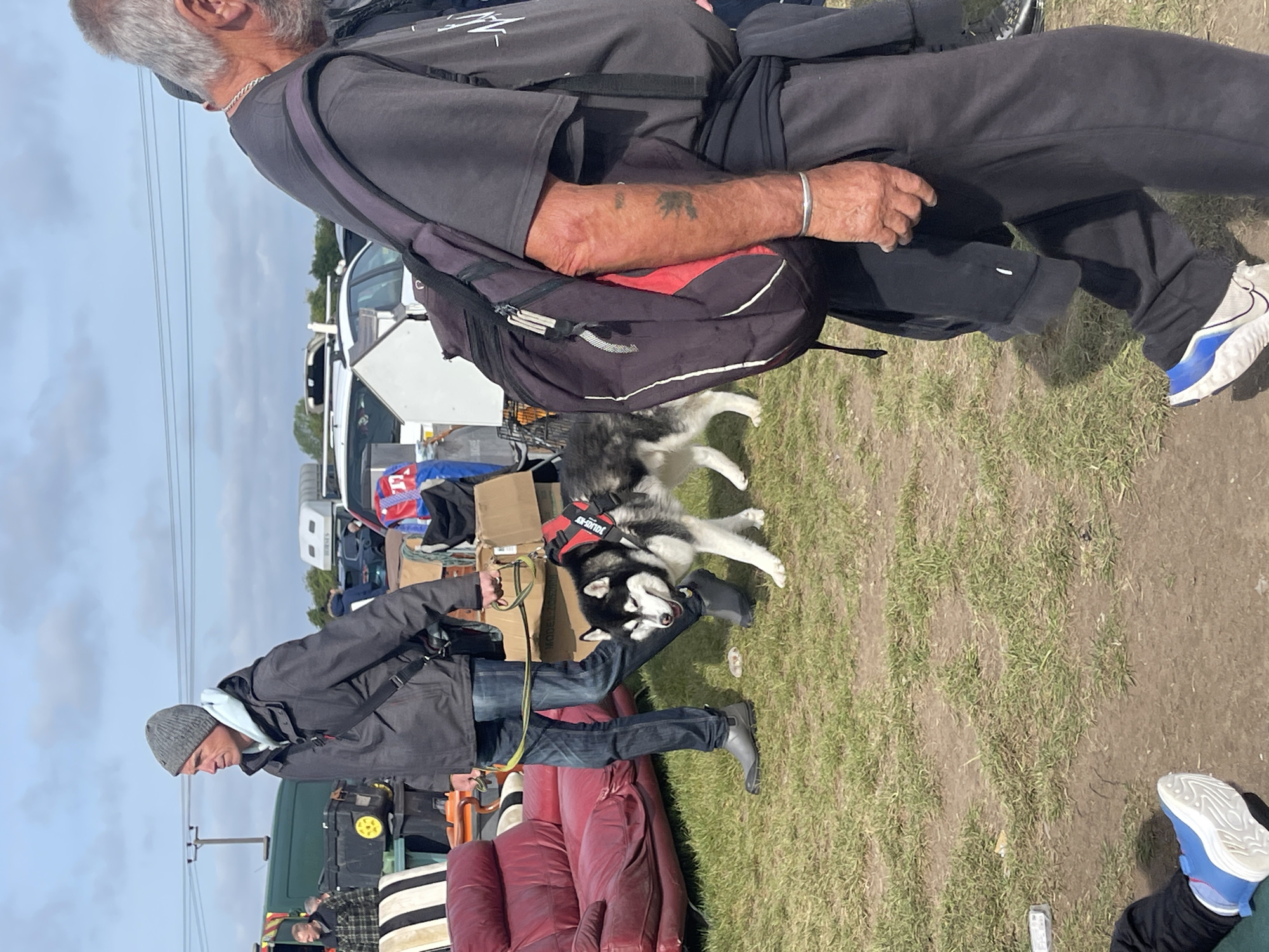 The image shows an outdoor scene, likely a field or grassy area, possibly in Sevenoaks, UK. There are several people present amidst a cluttered environment suggestive of a flea market or car boot sale. Two men are visible. The older man wears dark clothing and a backpack. He has grey hair and appears to be walking. A younger man is standing in front of a red couch; he wears a grey hat, jacket, and jeans. He is holding the leash of one or two huskies who appear to be excited. Vehicles, including vans and cars, are parked nearby, and various objects such as boxes, furniture, and assorted items are scattered around. The sky is overcast. Power lines are visible.