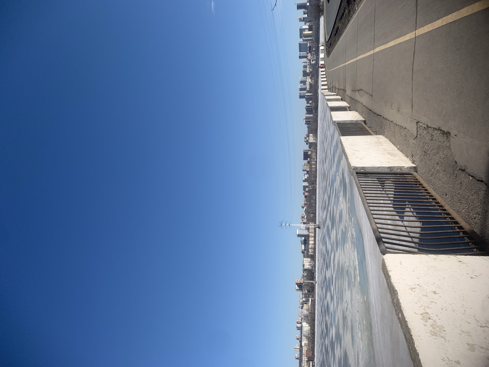 The image shows a road and a body of water with buildings in the background. The road has a yellow dividing line and is adjacent to a concrete barrier with metal grates. The body of water appears to be a river or lake, and reflections of the buildings are visible on the water's surface. The sky is clear and blue. Power lines are visible in the sky over the city. The location is Izhevsk, Russia, based on the architecture of the buildings.