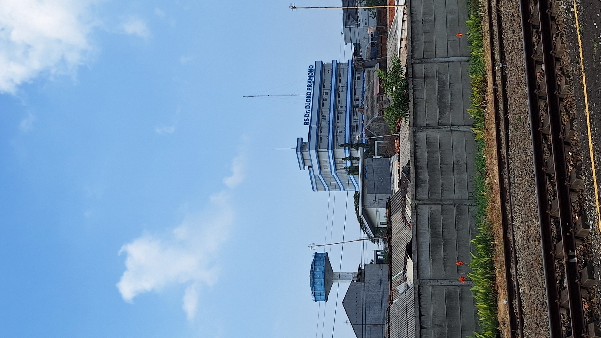 The image shows a cityscape with a large, multi-story building labeled "RS DR. DJOKO PRAMONO" prominently displayed. This building appears to be a hospital or a similar institution, featuring white walls with blue accents on its upper floors and balconies. To the left of this building, a tall water tower with the word "KARAWANG" written on it is visible. The surrounding environment consists of various structures with grey tiled roofs, a concrete wall, and lush green vegetation. In the foreground, railroad tracks are laid across a gravel bed. The sky is a clear blue with scattered white clouds. The scene is captured on a sunny day, with the lighting suggesting it is daytime. There are no people visible in the frame. The presence of the water tower with "KARAWANG" indicates the location is likely within or near the city of Karawang, Indonesia.