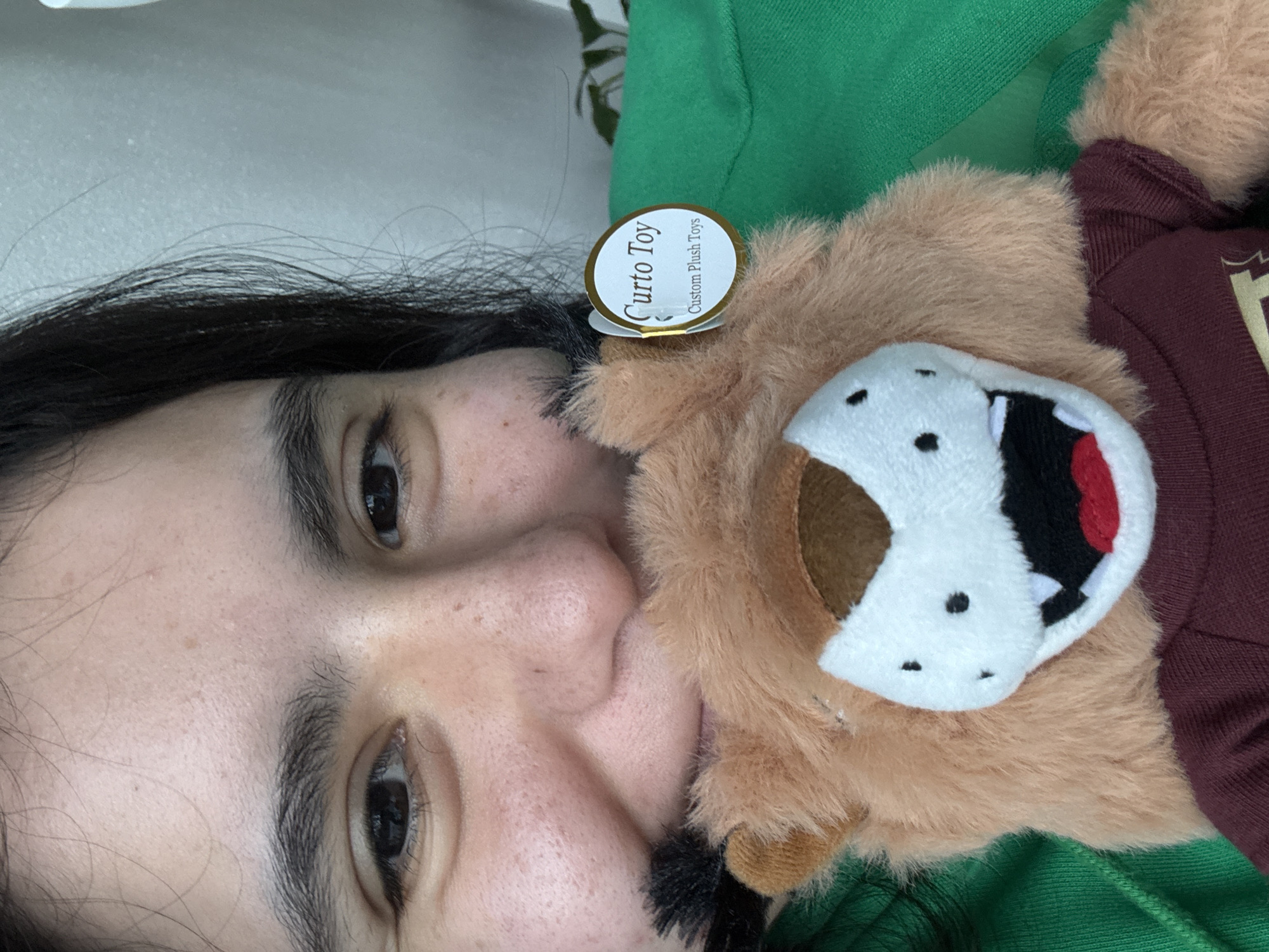 The image is a close-up shot of a person's face next to a plush toy. The person has dark hair and is looking towards the camera. The plush toy appears to be a lion mascot of some kind, wearing a burgundy-colored top. A tag is visible on the plush toy that reads "Curto Toy, Custom Plush Toys." The person is wearing a green shirt. It is not possible to determine the location in Austin, Texas, from the image.