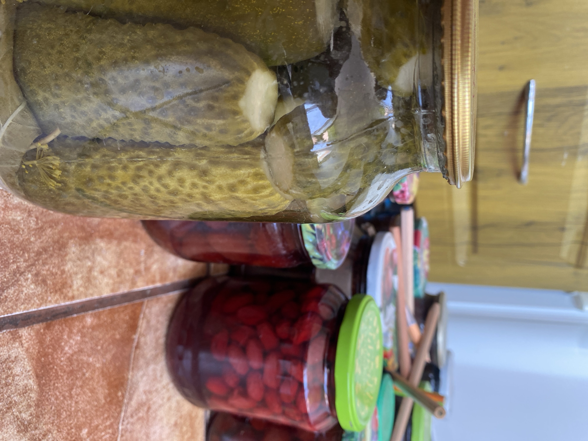 In a close-up shot, a large glass jar filled with preserved cucumbers takes center stage, positioned on a tiled surface. The jar is sealed with a golden lid. Behind it, several other glass jars are visible, containing a dark red preserve, possibly berries or jam, and some jars with red beans. The background features a wooden cabinet and a white appliance, suggesting an indoor setting. The scene appears to be a home kitchen or pantry. The image was captured in Chornomorsk, Ukraine.