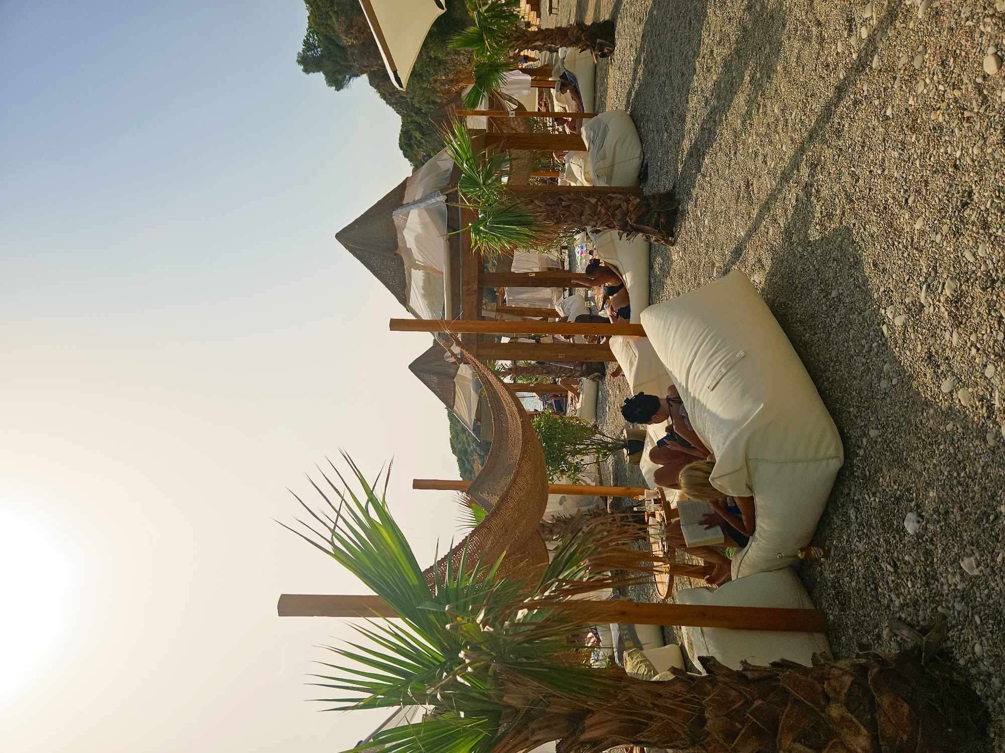The image depicts a beach resort with several people relaxing on beanbag chairs under thatched-roof cabanas. Palm trees are visible, and the ground is covered in pebbles. The scene is set on a sunny day, with clear skies and warm lighting. In the foreground, a woman reads a book while another person lies next to her on a beanbag. A small table with drinks sits between them. In the background, more cabanas and people can be seen enjoying the beach. The resort appears to be located in a scenic coastal area, possibly with cliffs or hills in the distance.